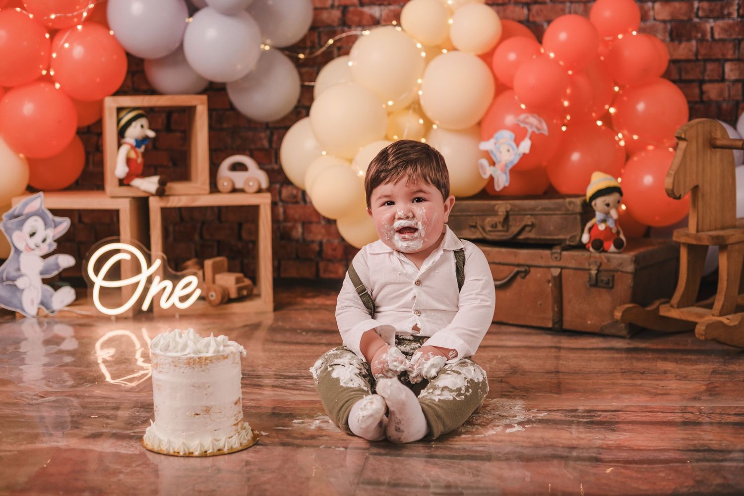 A baby's first birthday cake smash photo session with red and white balloons against a rustic brick wall backdrop.