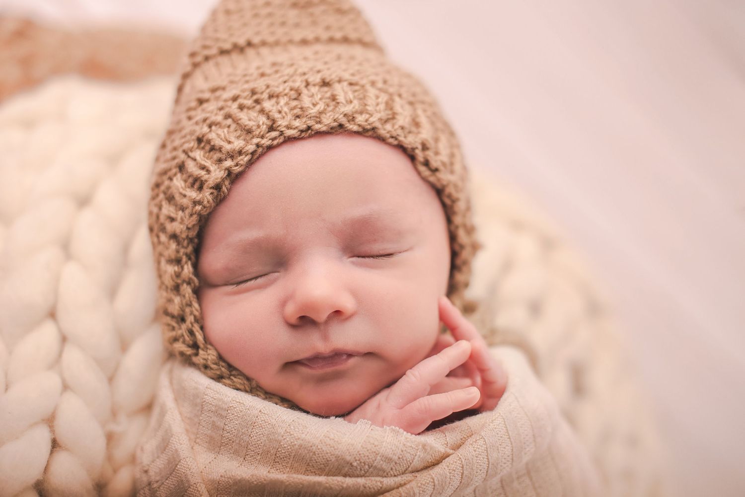 Peaceful sleeping newborn wearing tan knit hat on cream background.