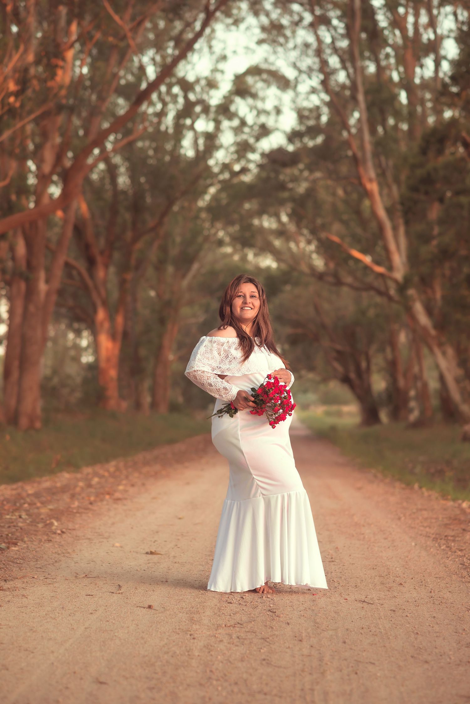 A person in a white dress holds red flowers while walking along a scenic dirt path through eucalyptus trees at sunset.