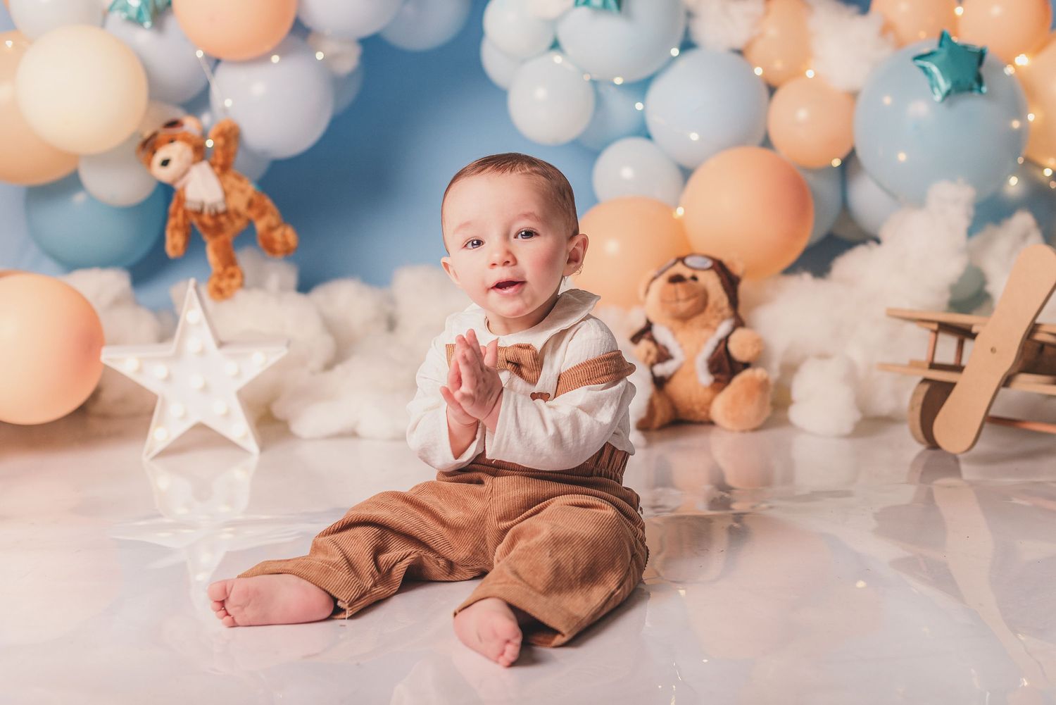 Baby in brown overalls sits on white backdrop with peach and blue balloons and star decorations.