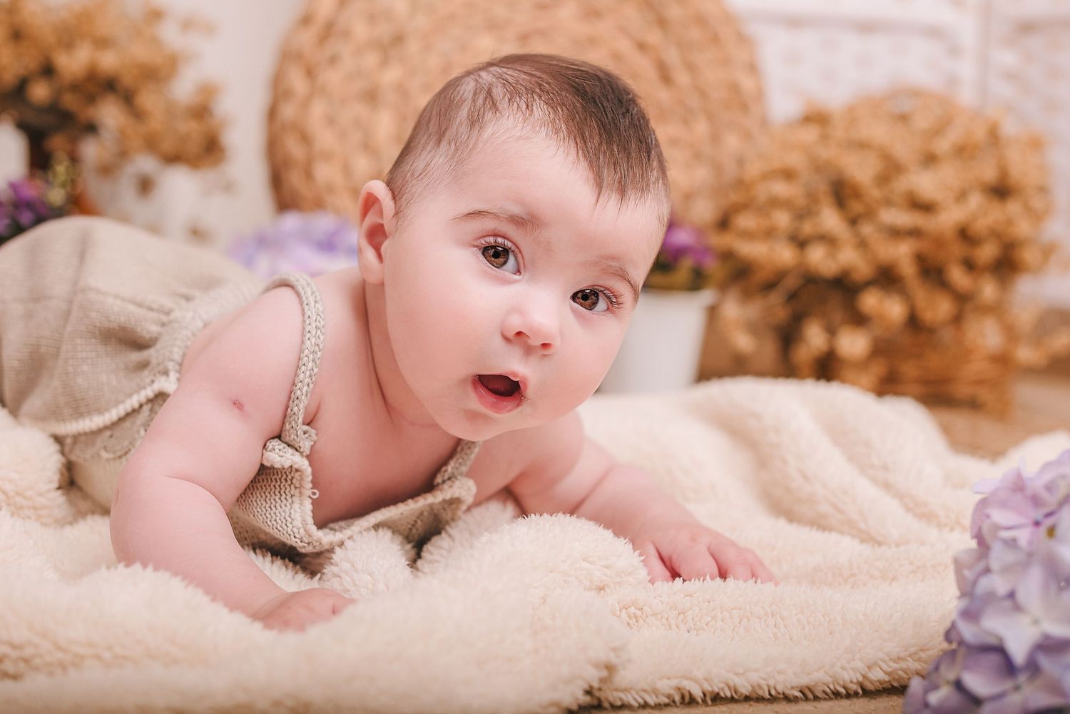 Baby lying on soft cream blanket with plush toys and purple flowers in background.