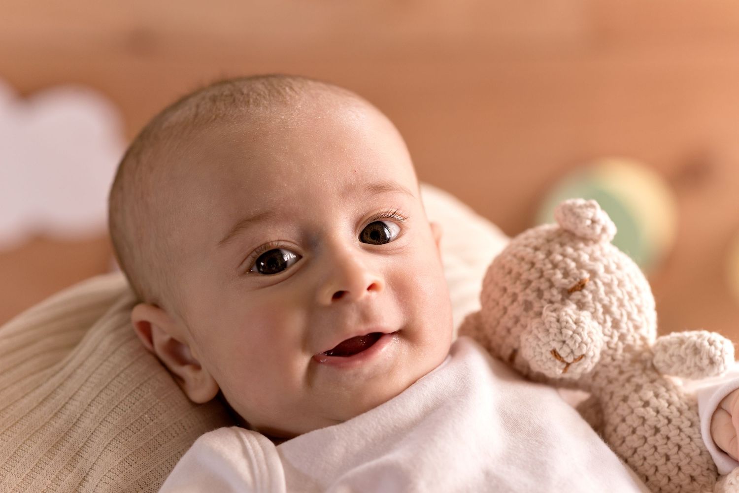 Baby lying on soft beige blanket with teddy bear smiles warmly at camera in natural light.