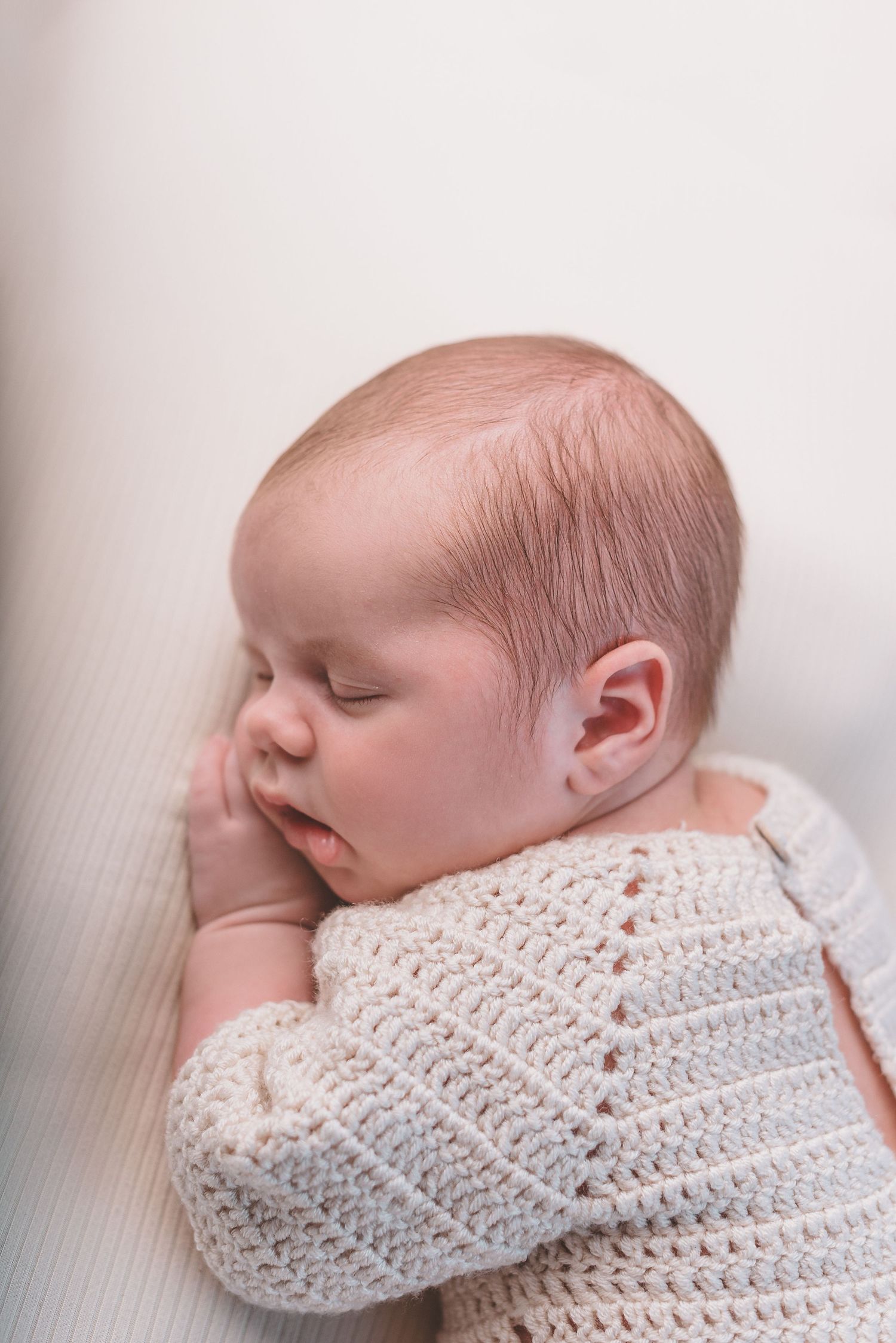 Sleeping baby wearing white knitted sweater rests peacefully against light background.