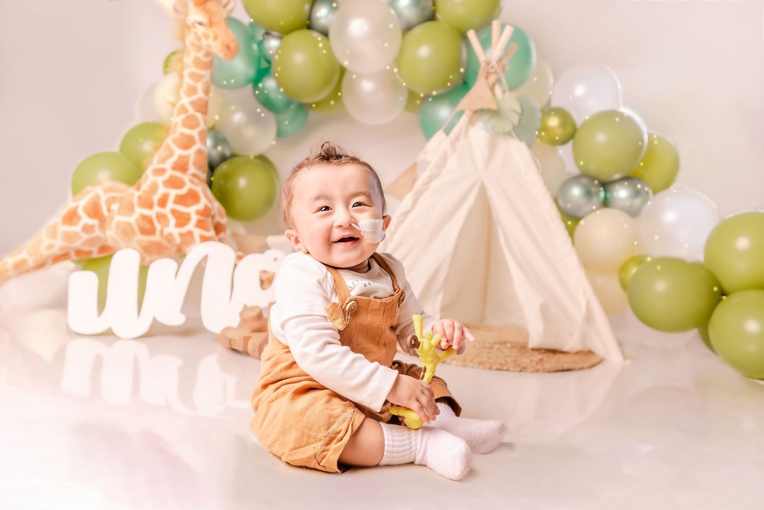 Baby sits on floor with giraffe decoration and green balloon arch for first birthday photoshoot setup.
