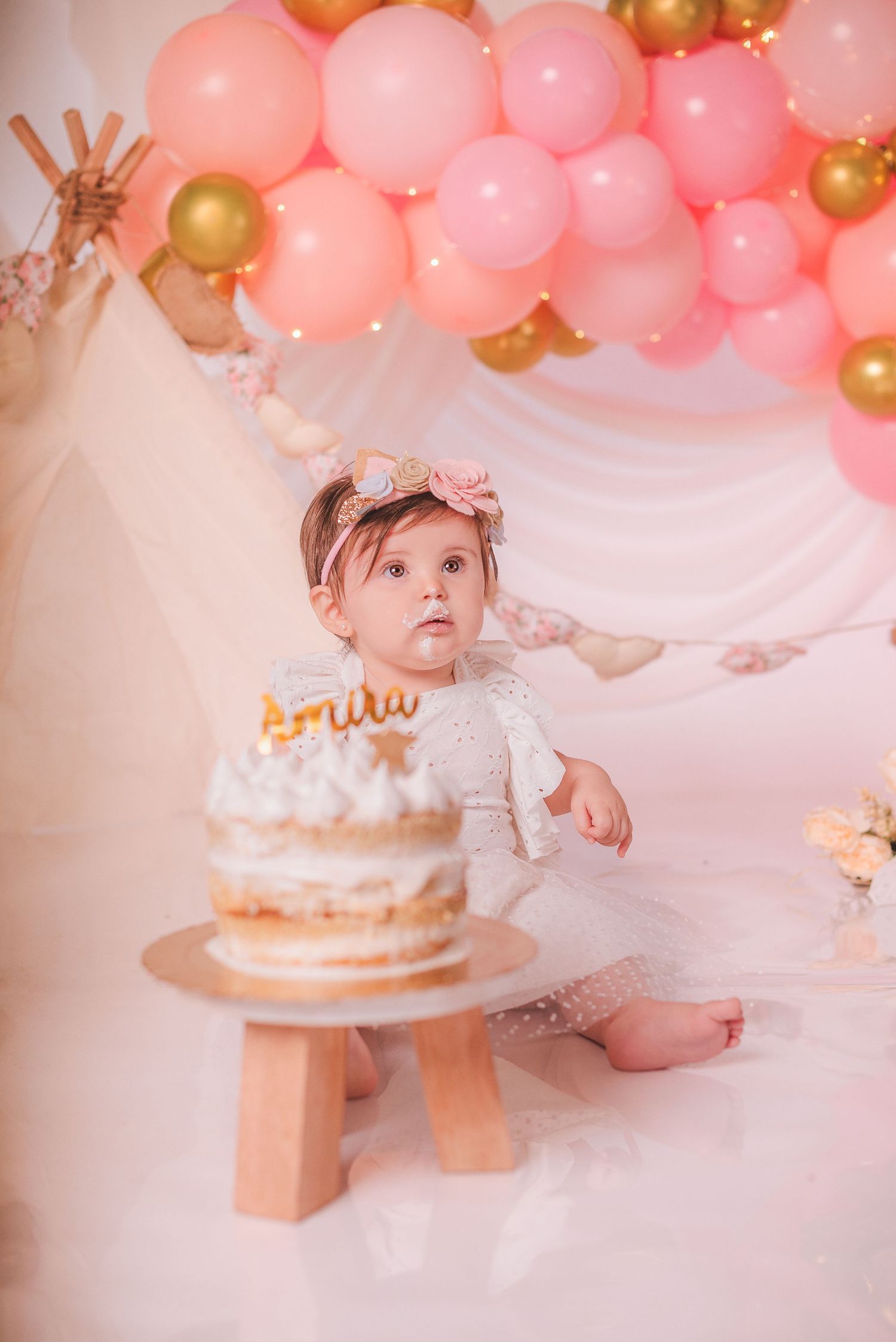 Pink and gold balloon garland decorates a first birthday cake smash photo session with soft dreamy lighting.