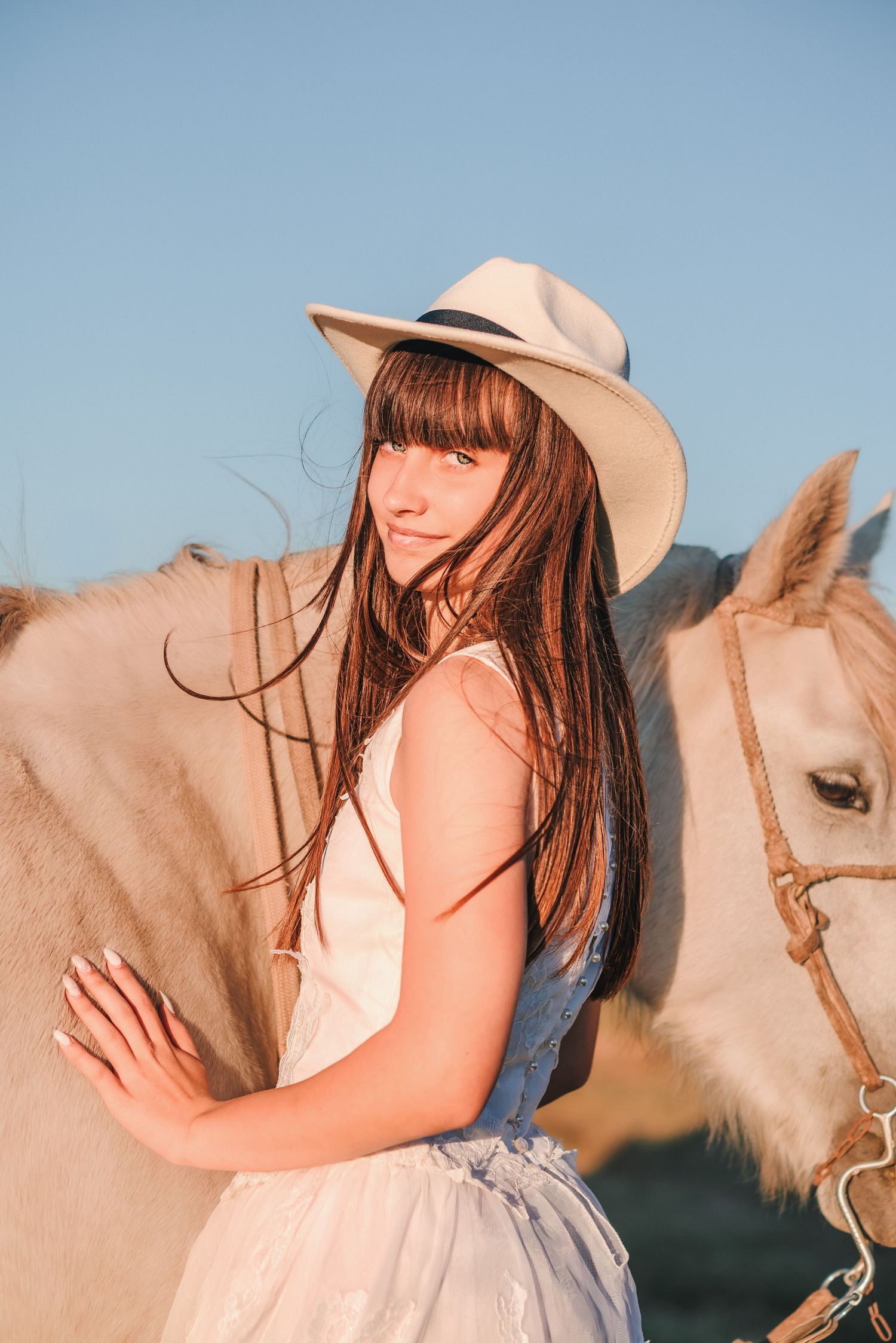 A woman in a cowboy hat and white dress stands affectionately next to a light-colored horse at sunset.