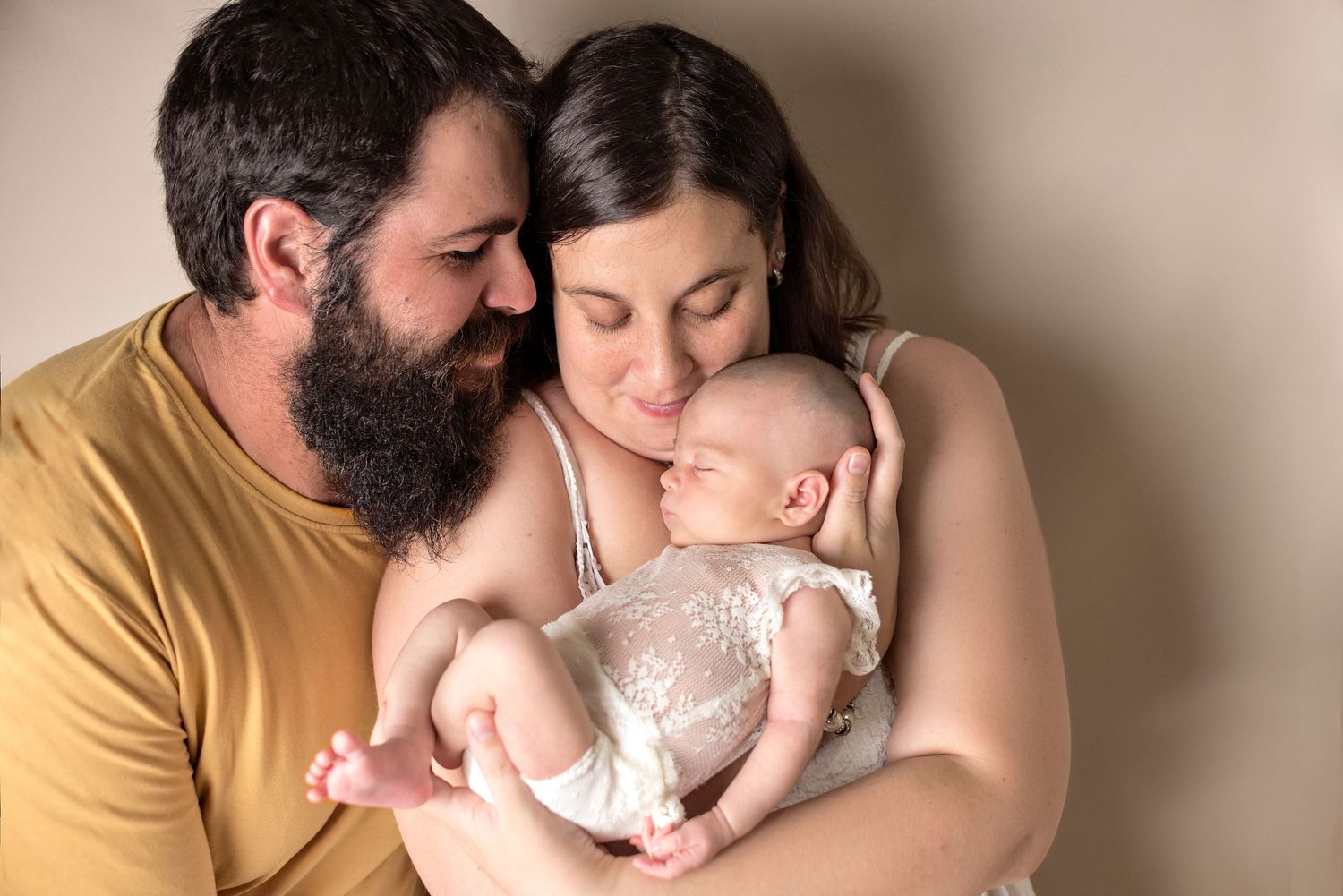 A tender family moment as parents embrace their sleeping newborn baby wearing white lace against neutral background.