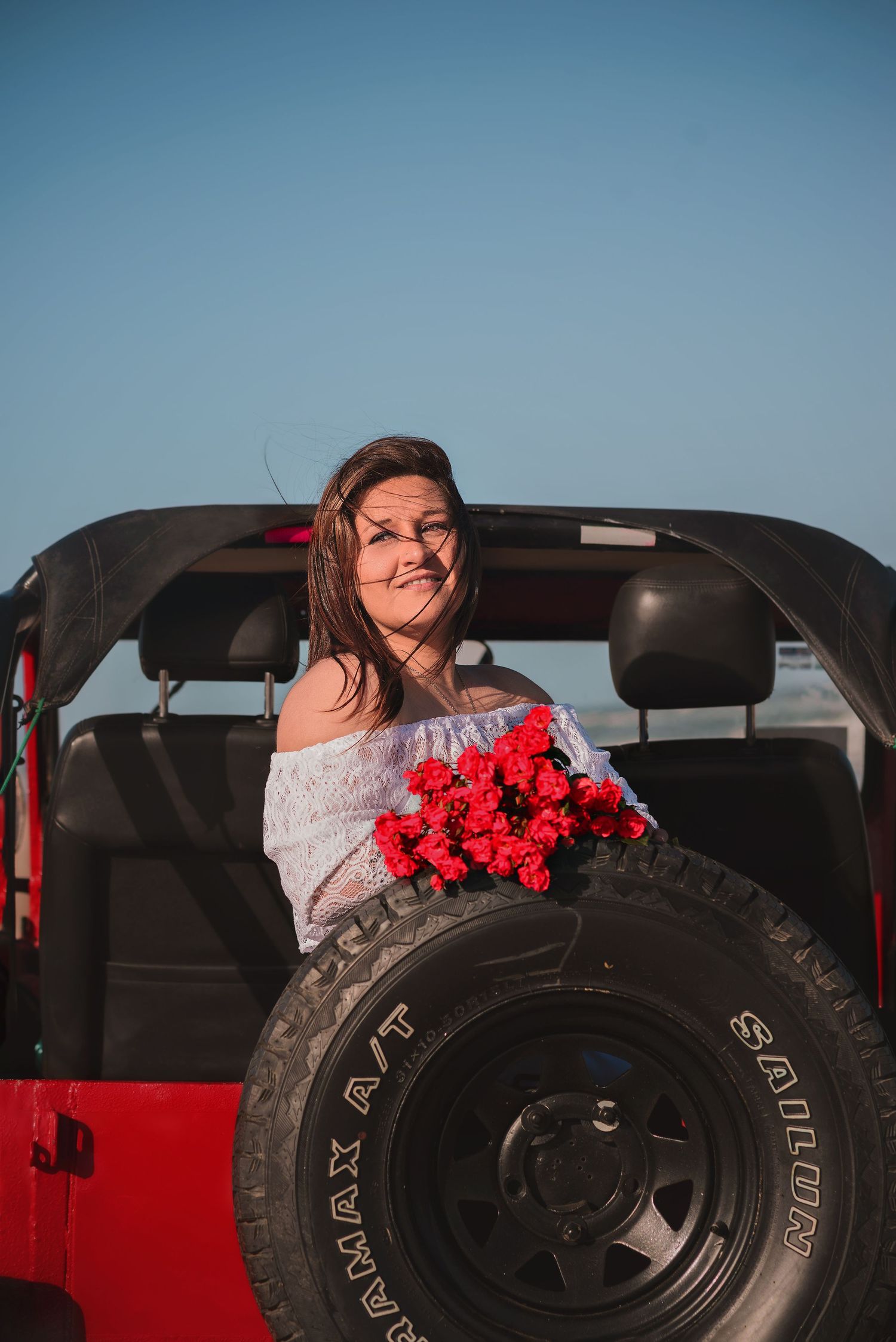 A person in a white off-shoulder top poses with red roses next to a black spare tire mounted on a red Jeep.