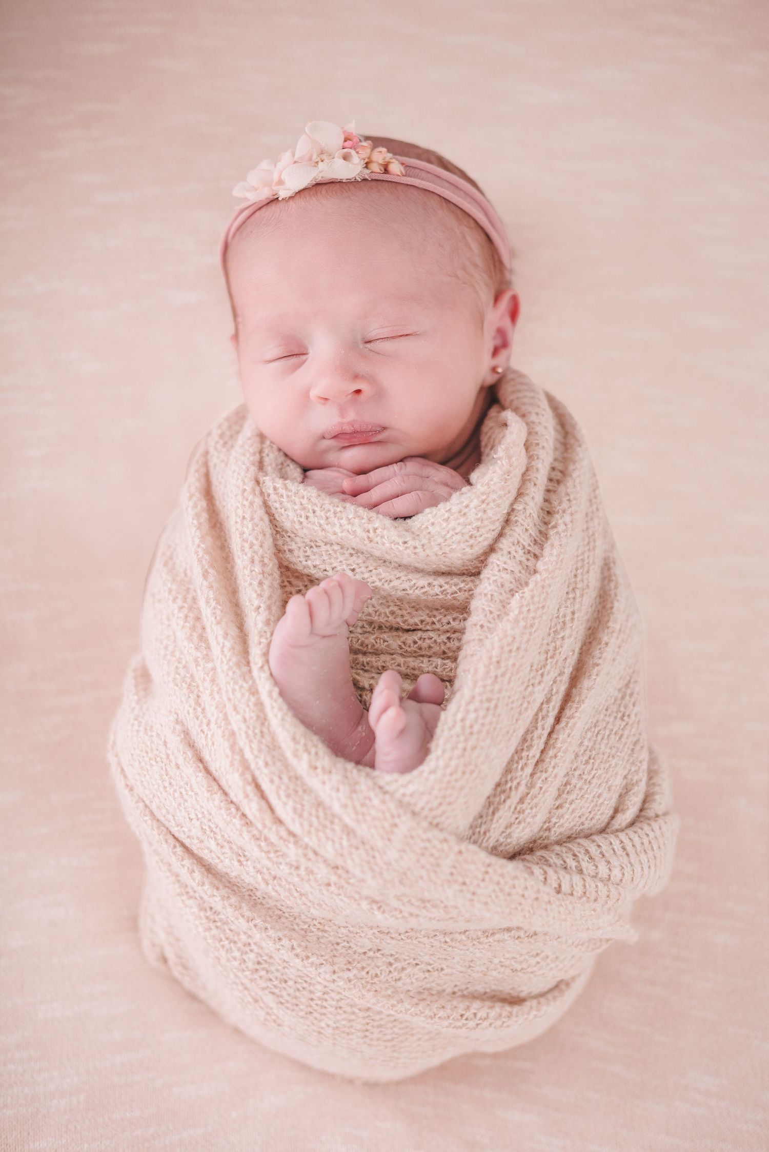 Sleeping newborn baby wrapped in soft beige knit blanket wearing delicate pink headband on pink background.
