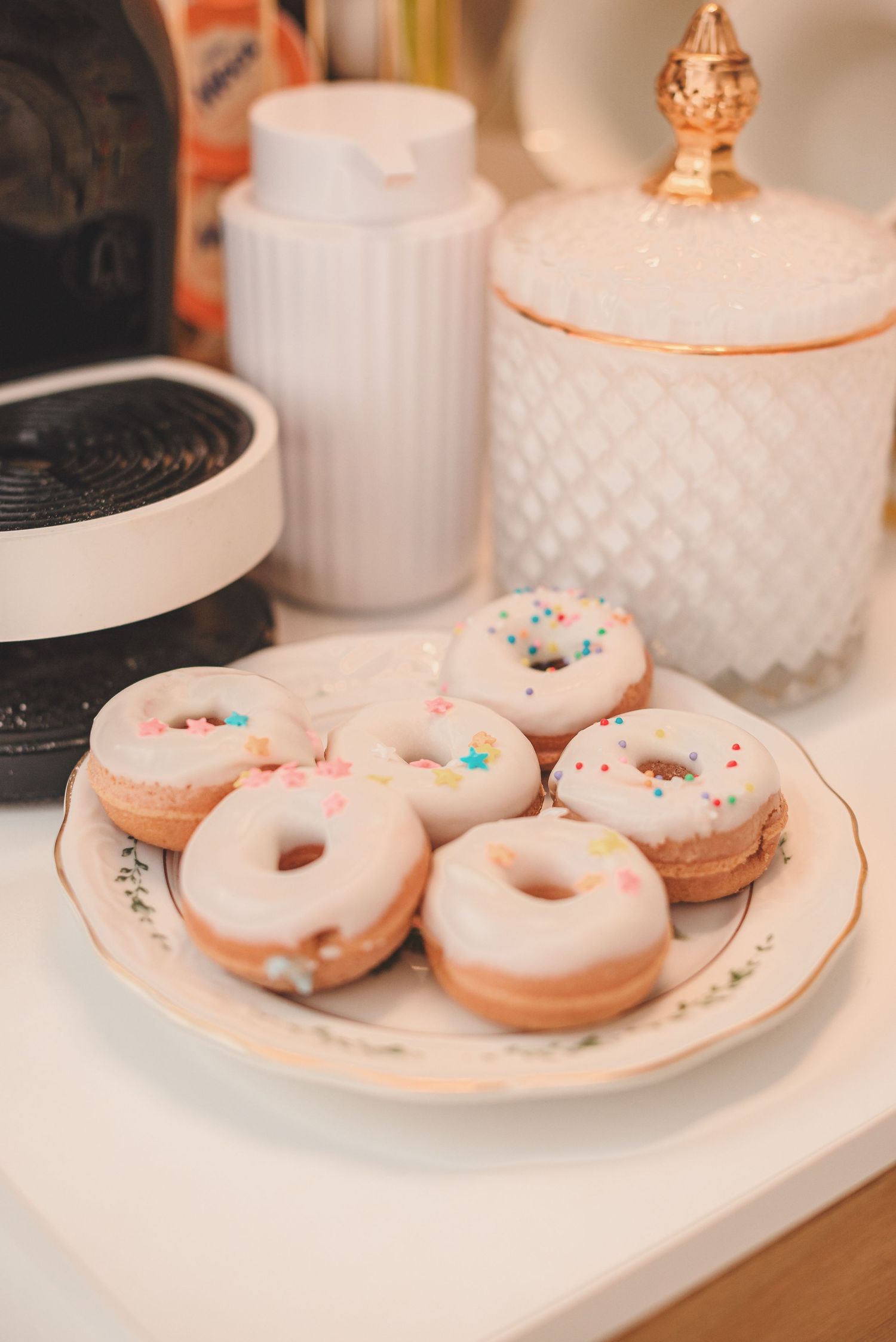 Glazed vanilla donuts with colorful sprinkles displayed on white plates against a marble countertop background.