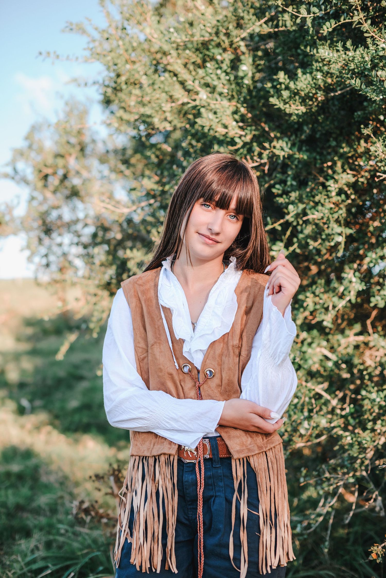 A person wearing a brown fringed vest and white blouse poses outdoors against a natural backdrop with trees.