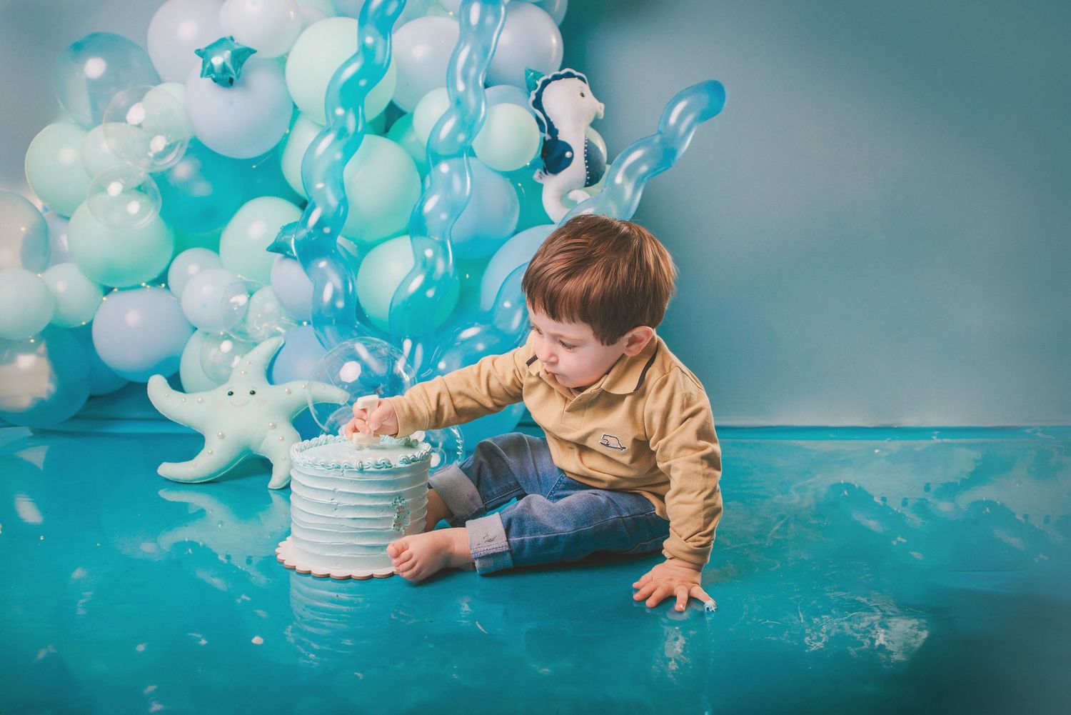 Baby in white dress explores blue and white balloon decorations during cake smash photo session in studio.