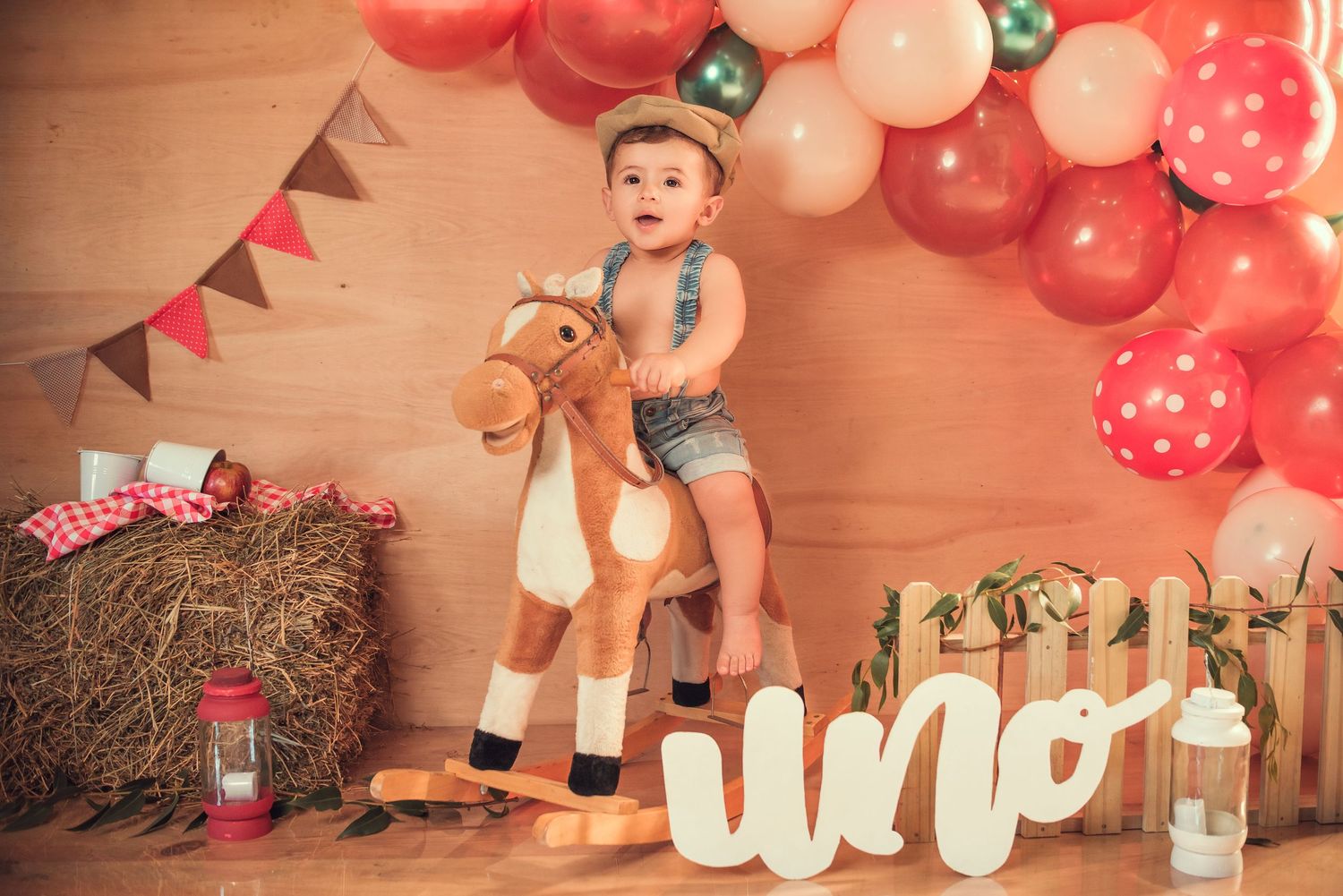A festive first birthday party setup with pink and coral balloons, wooden decorations, and rustic farmhouse elements.