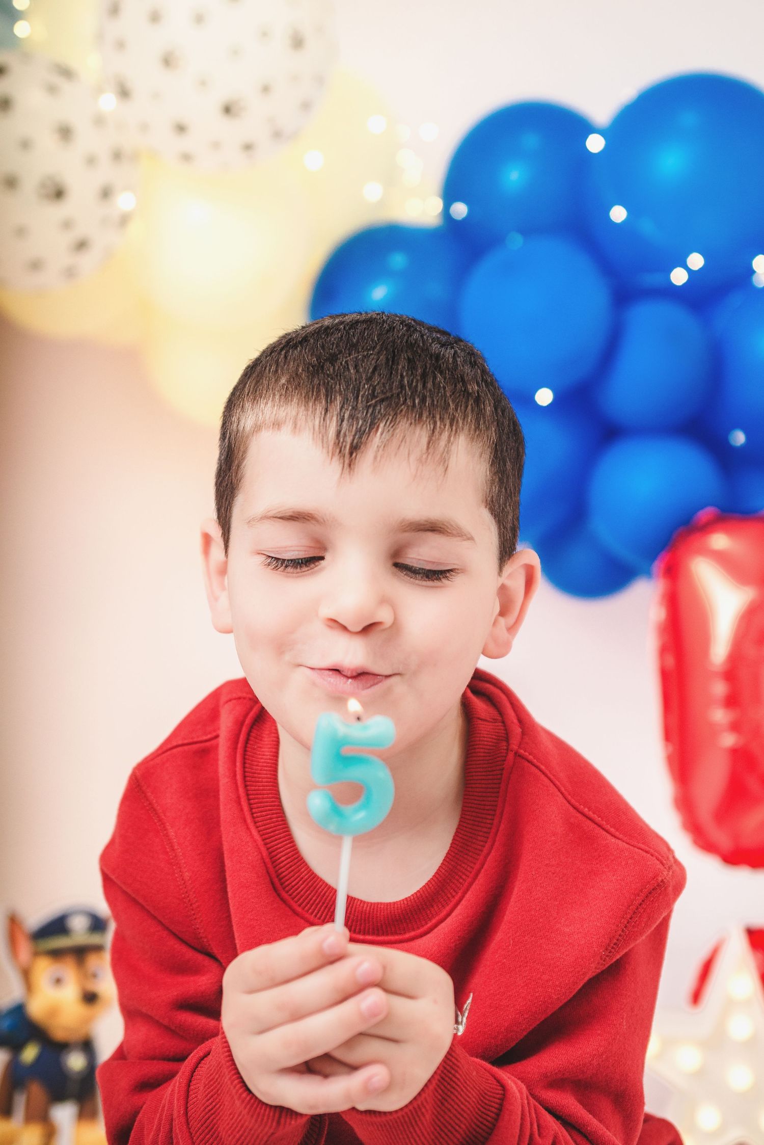 A child in a red sweatshirt holds a blue number 5 candle with birthday balloons in the background.