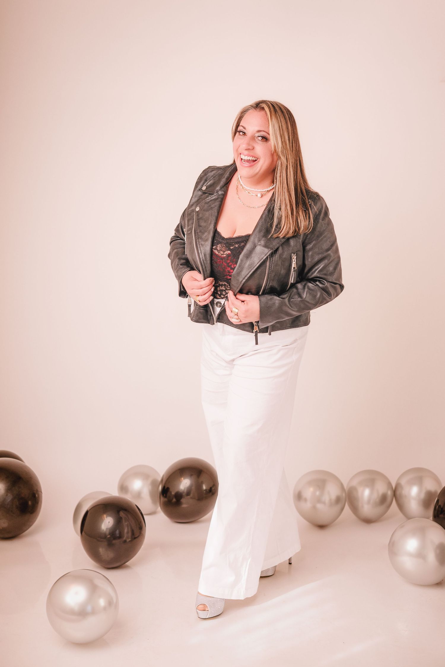 A person in a leather jacket and white dress poses with silver and brown balloons against a light pink backdrop.