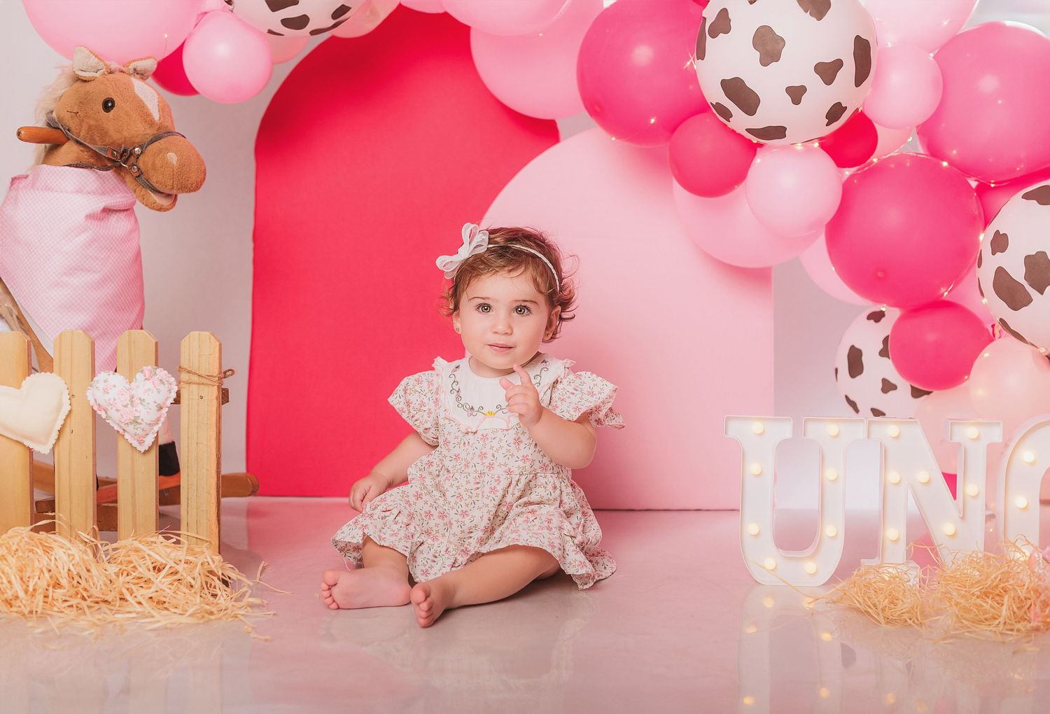Pink and white balloon arch decorates a first birthday photoshoot setup with rustic wooden fence and straw accents.