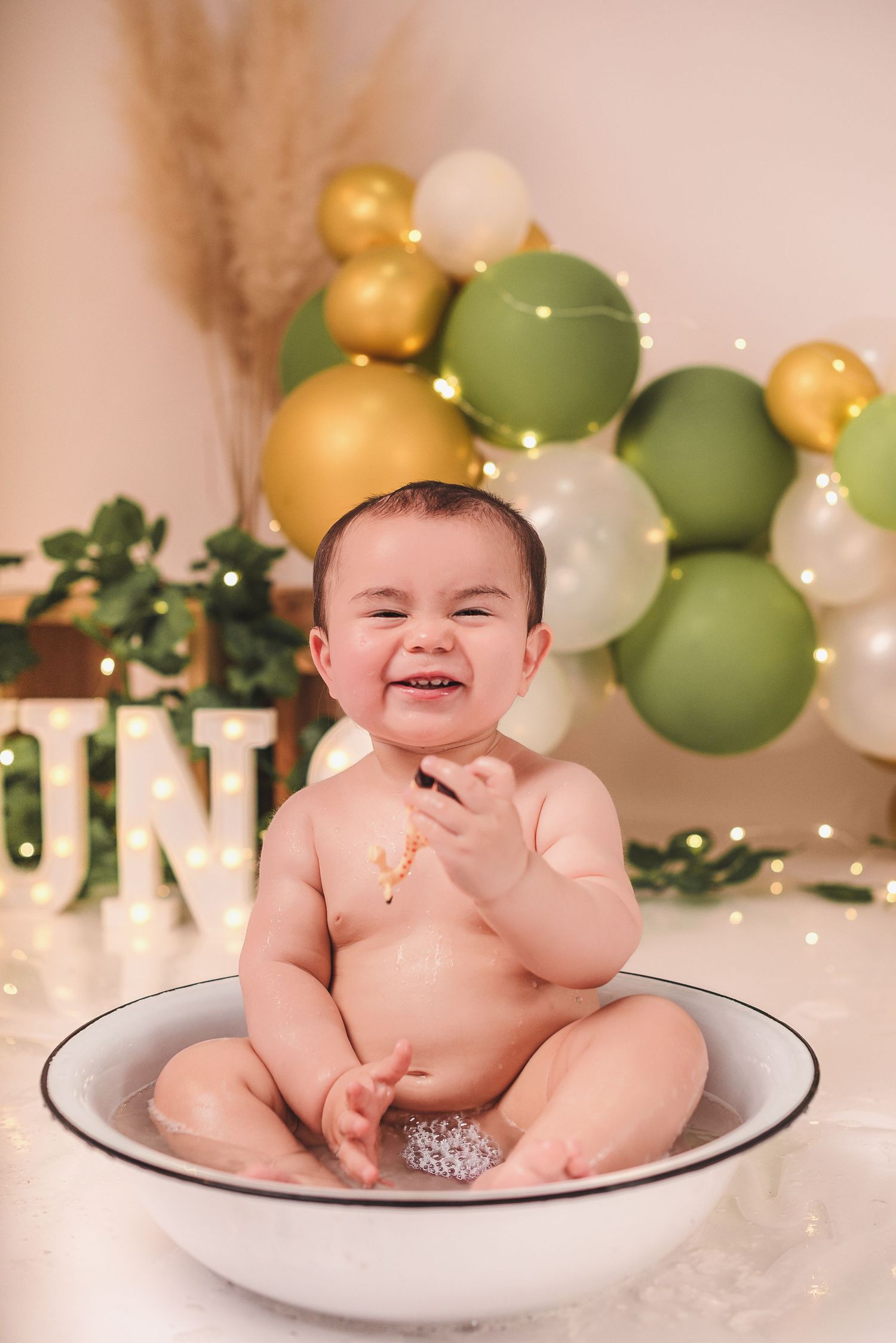 Baby sits in white enamel basin surrounded by green and gold balloons for milestone photo session.