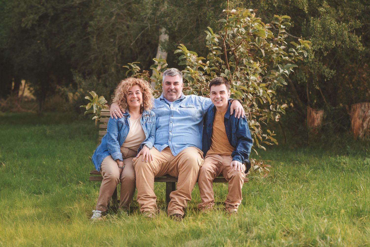 Three people sit together on a rustic wooden bench in a sunny garden setting with green grass and trees.