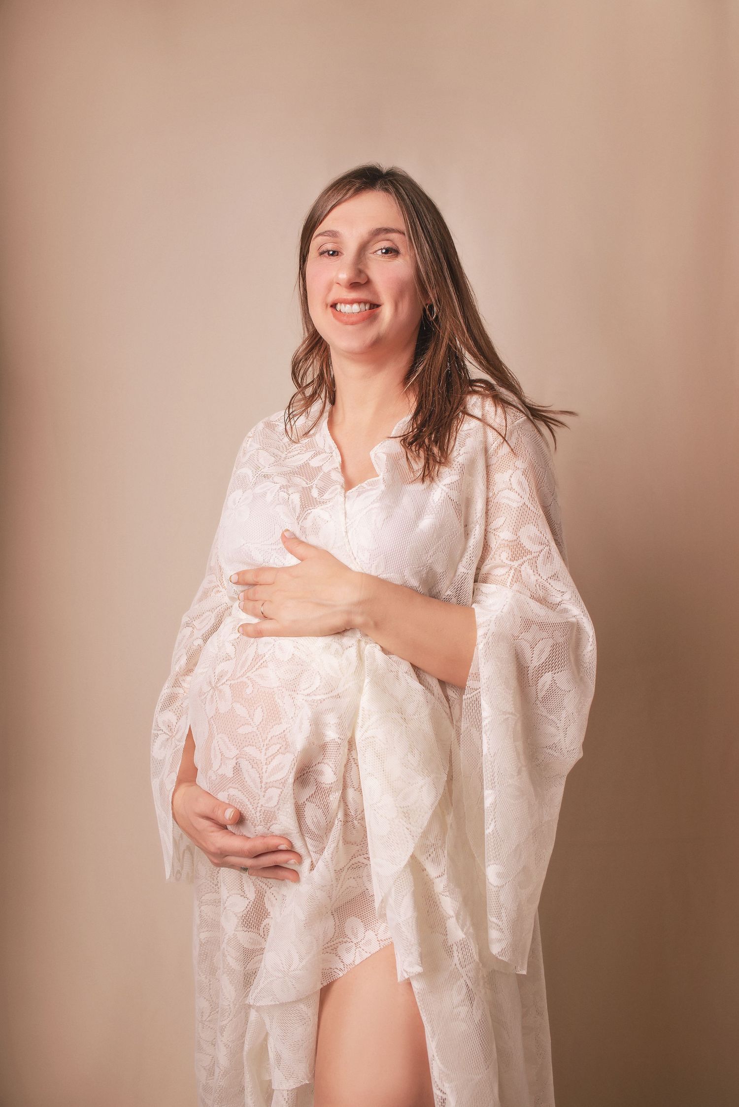 Maternity portrait with flowing lace robe against neutral background.