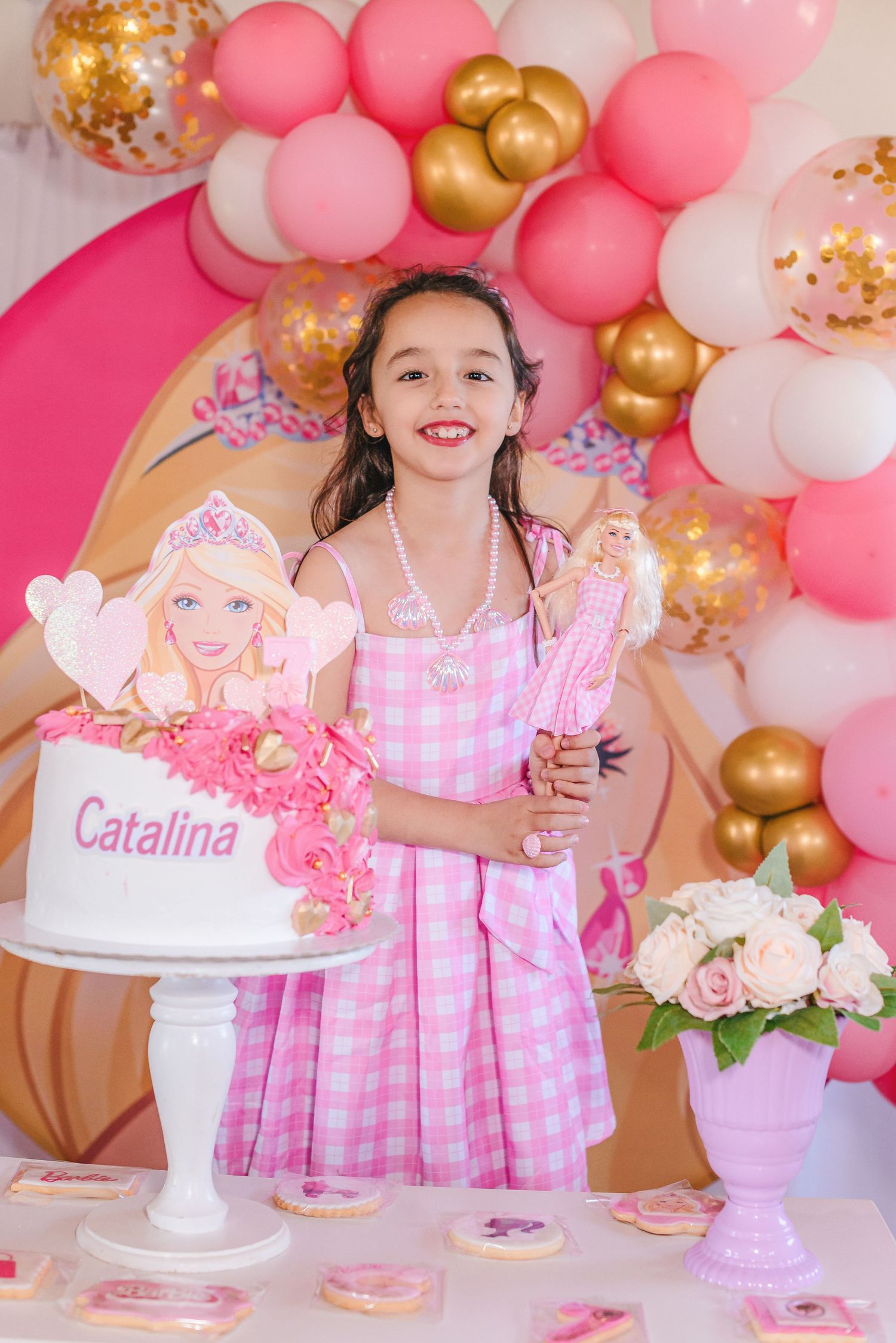 Pink and gold balloon garland decorates a festive party setup with cake stands and gingham dresses in soft pastel colors.