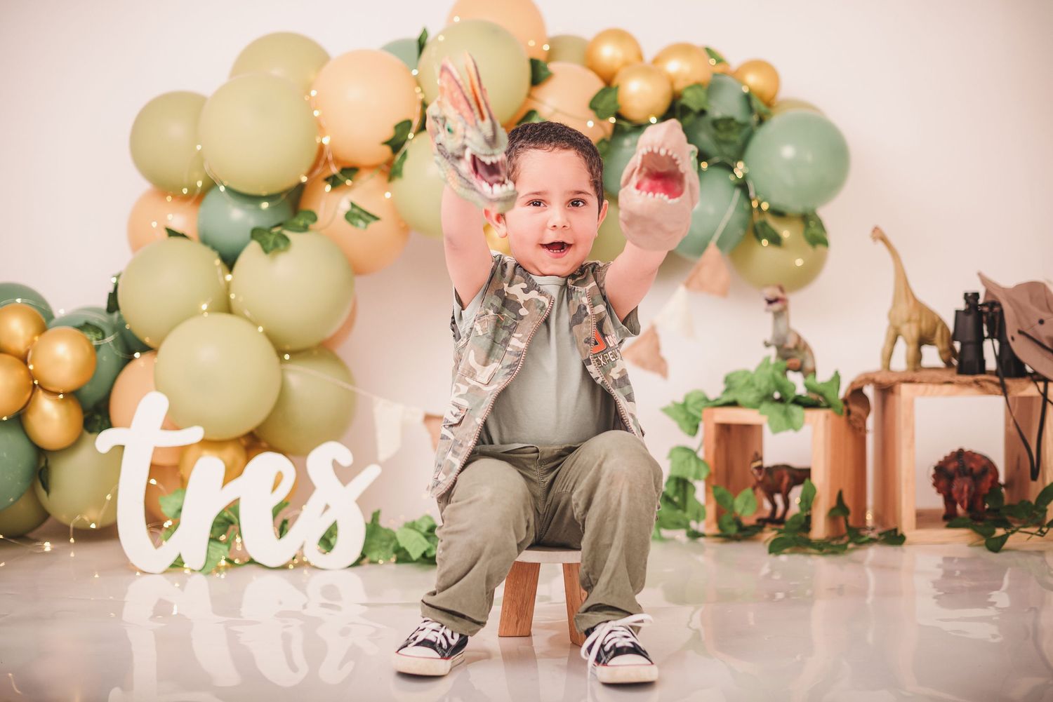 A festive birthday setup with sage green and gold balloon arch, decorative 'tres' sign, and safari-themed party decorations.