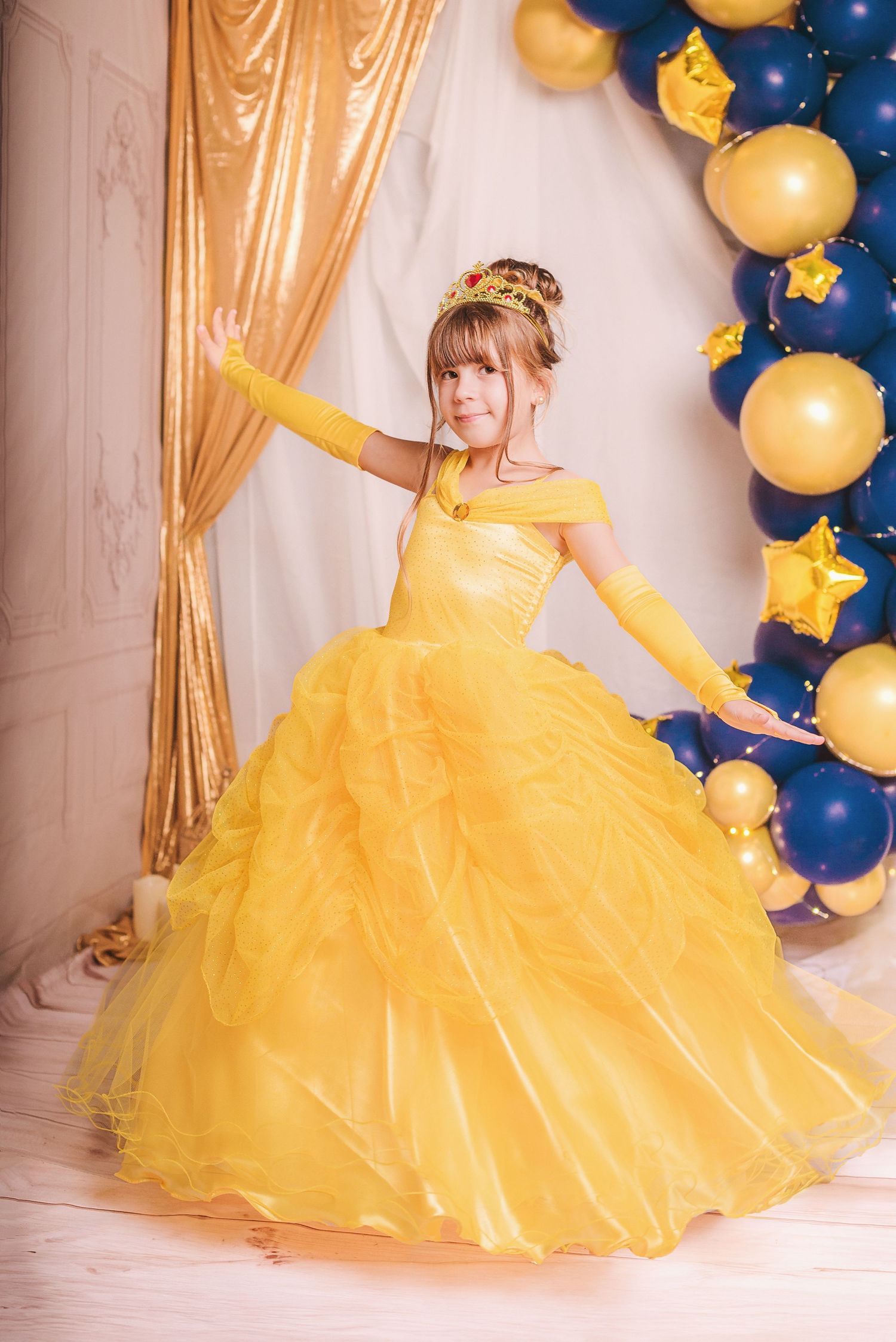 A flowing yellow princess ball gown with tulle skirt twirls against a backdrop of navy blue and gold balloon decorations.