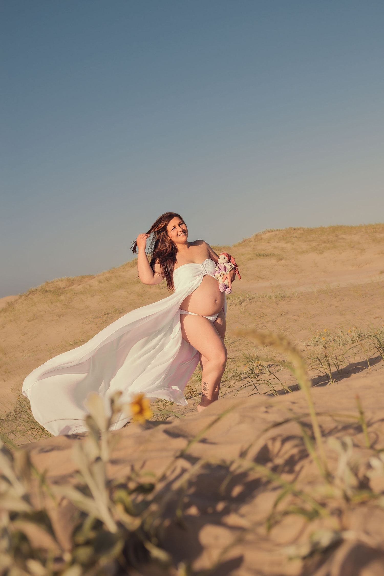 A flowing white dress billows in the desert wind against sandy hills and clear blue sky at sunset.