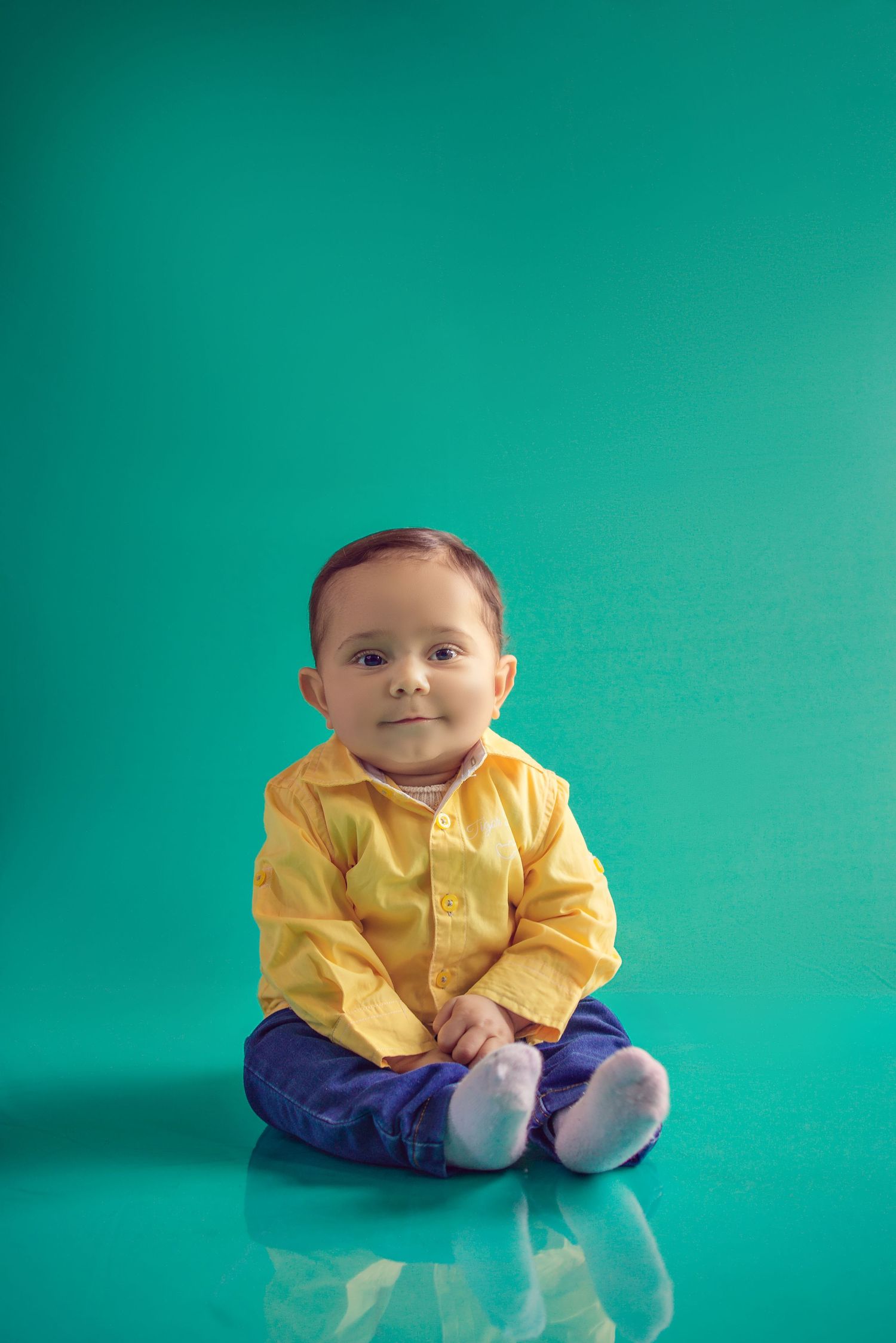 Baby wearing yellow shirt and purple pants sits on reflective surface against turquoise background.