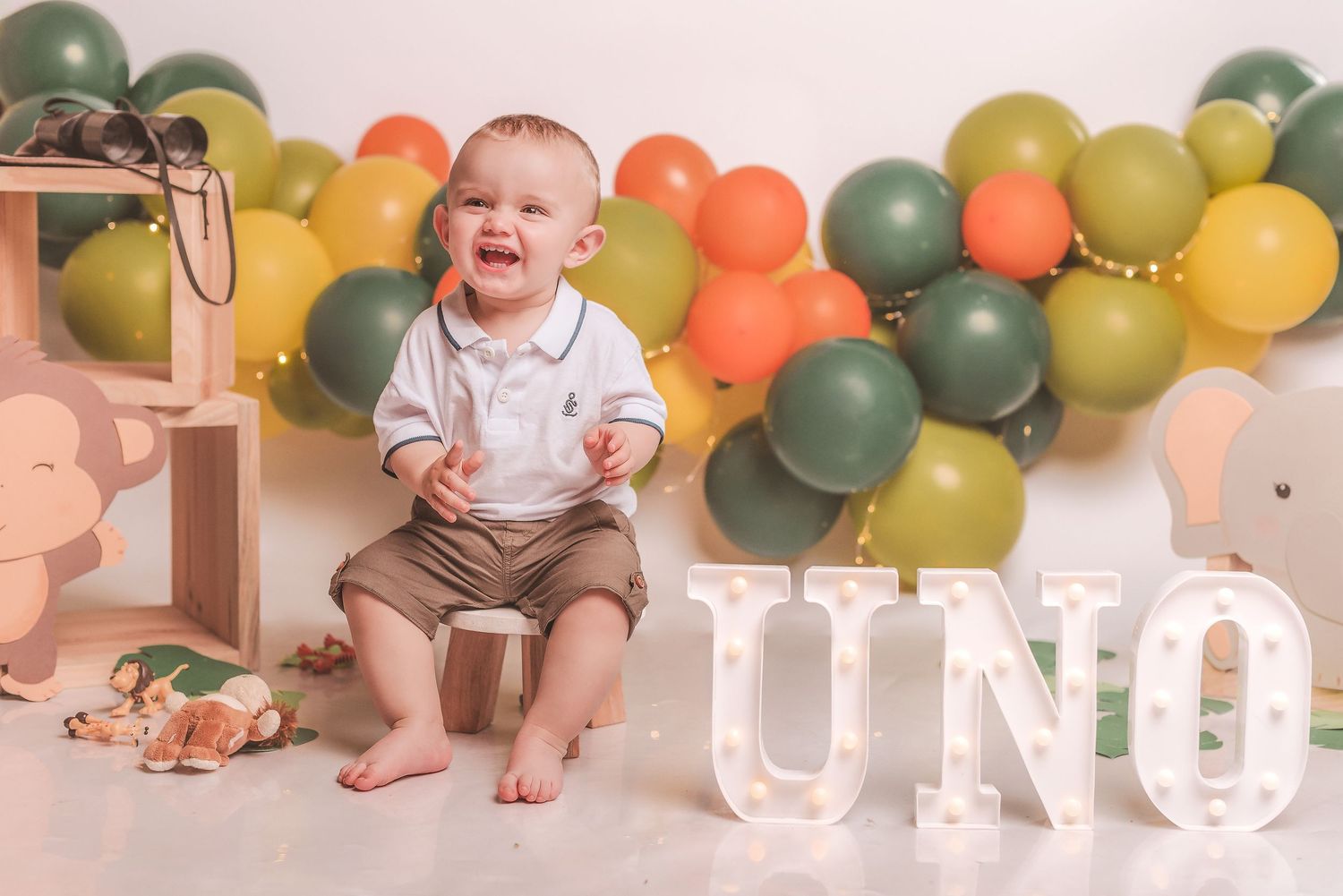 A festive first birthday party setup with green and yellow balloons and a smiling baby in white clothing.