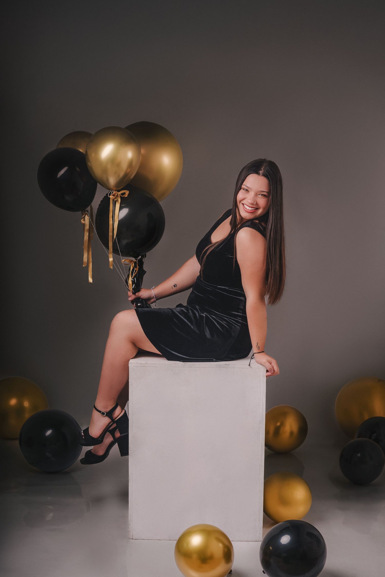 A person in a black dress poses with gold and black balloons against a gray backdrop in a glamorous photoshoot.