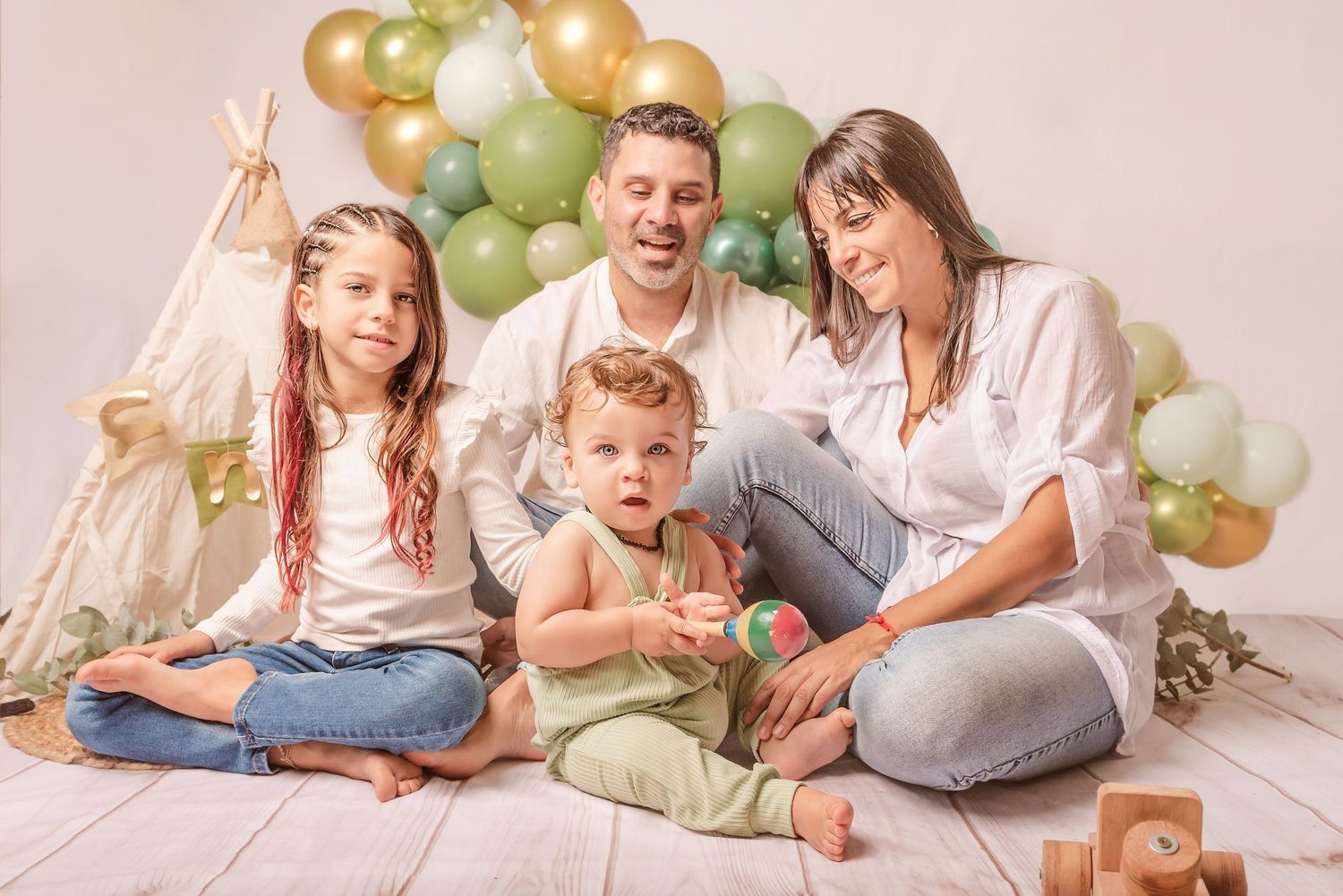 Family sitting on floor with festive green and gold balloon arch decoration in pastel studio setting.