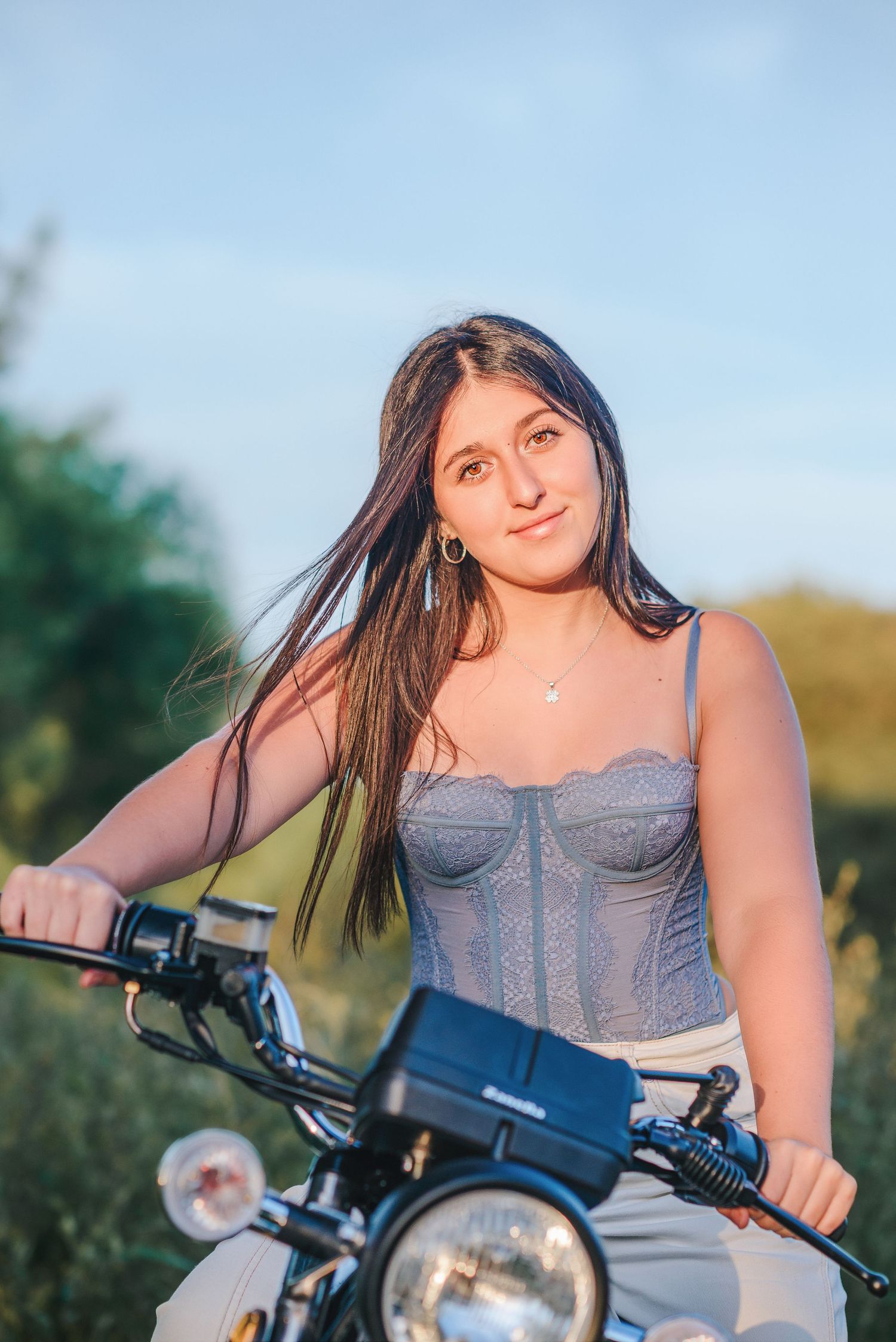 A person sits on a motorcycle wearing a gray corset top against a blurred natural background during golden hour.