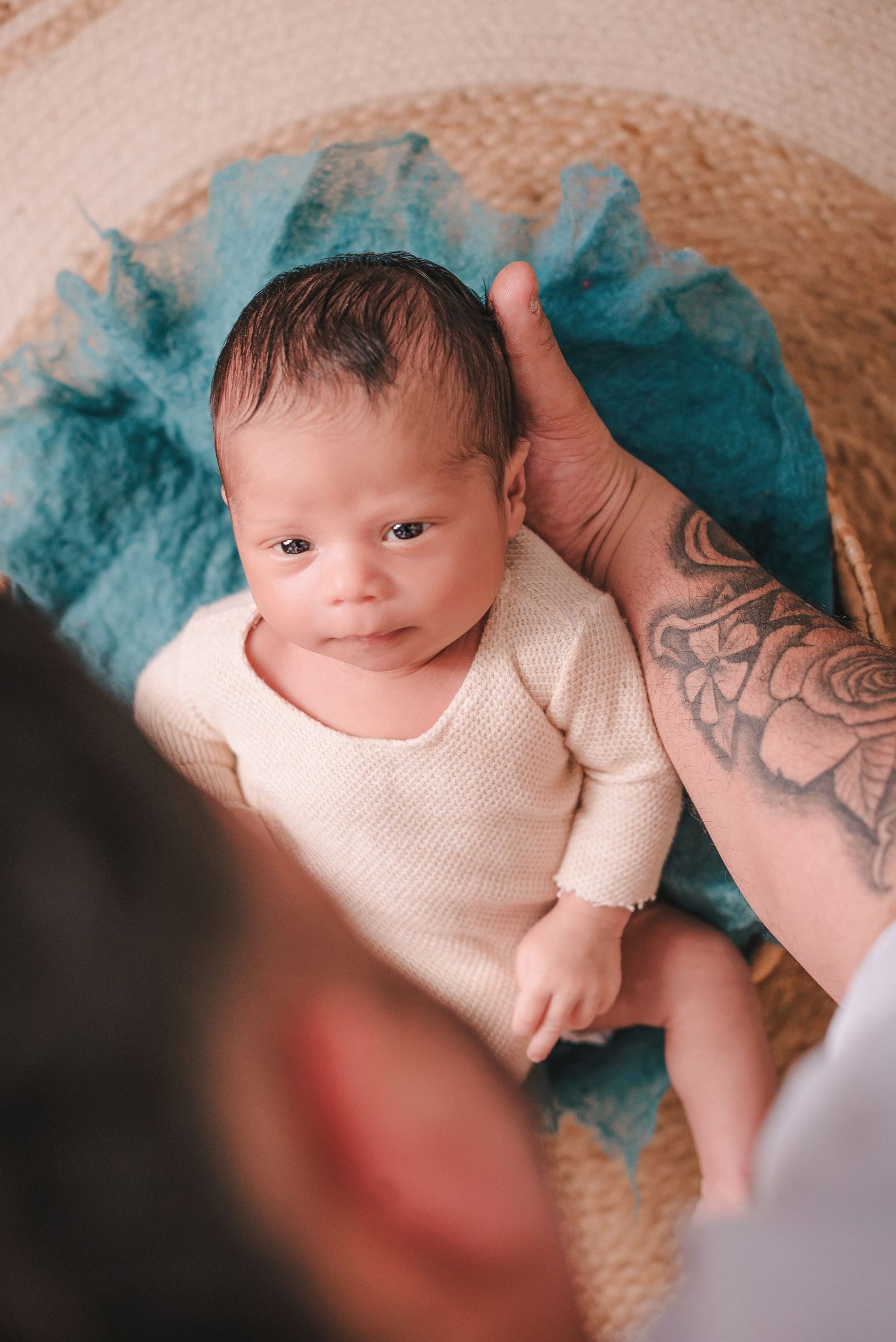 A small baby wearing a cream sweater lies on turquoise tulle fabric while a tattooed arm reaches into frame.