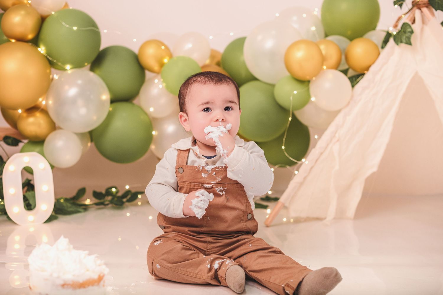 Baby in brown outfit sits on floor surrounded by green and gold balloon arch at special celebration event.
