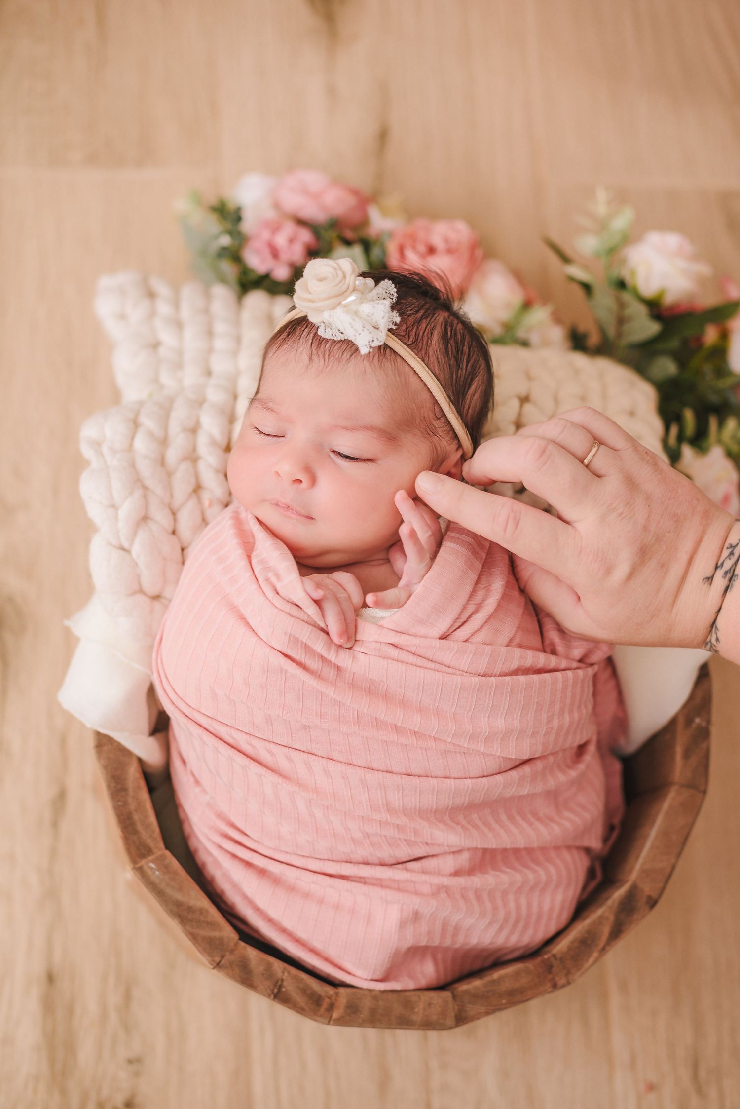 A sleeping newborn wrapped in soft pink fabric lies peacefully in a rustic wooden bowl with floral decorations.