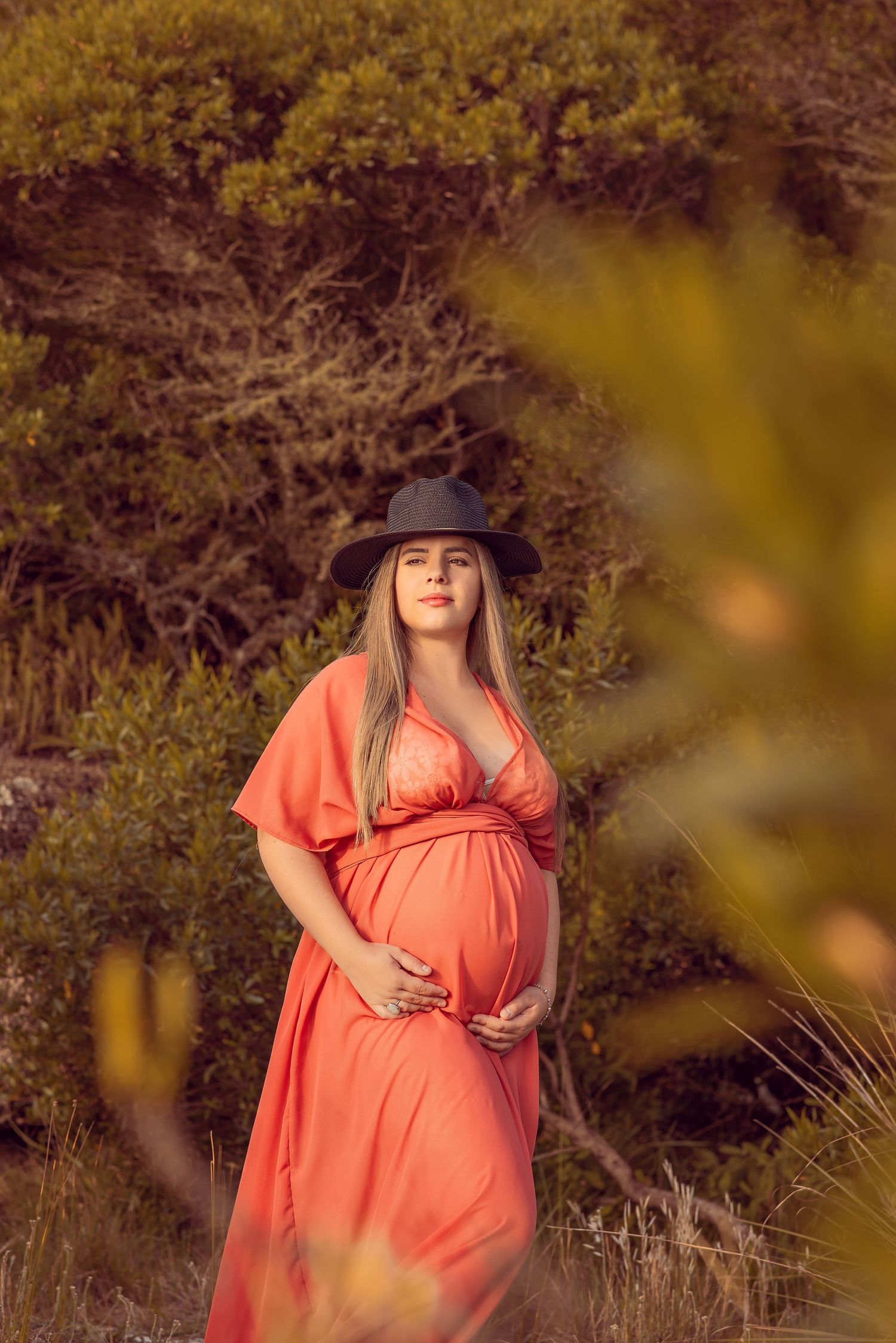 A person in a coral dress and wide-brimmed hat poses outdoors during golden hour among autumn foliage.