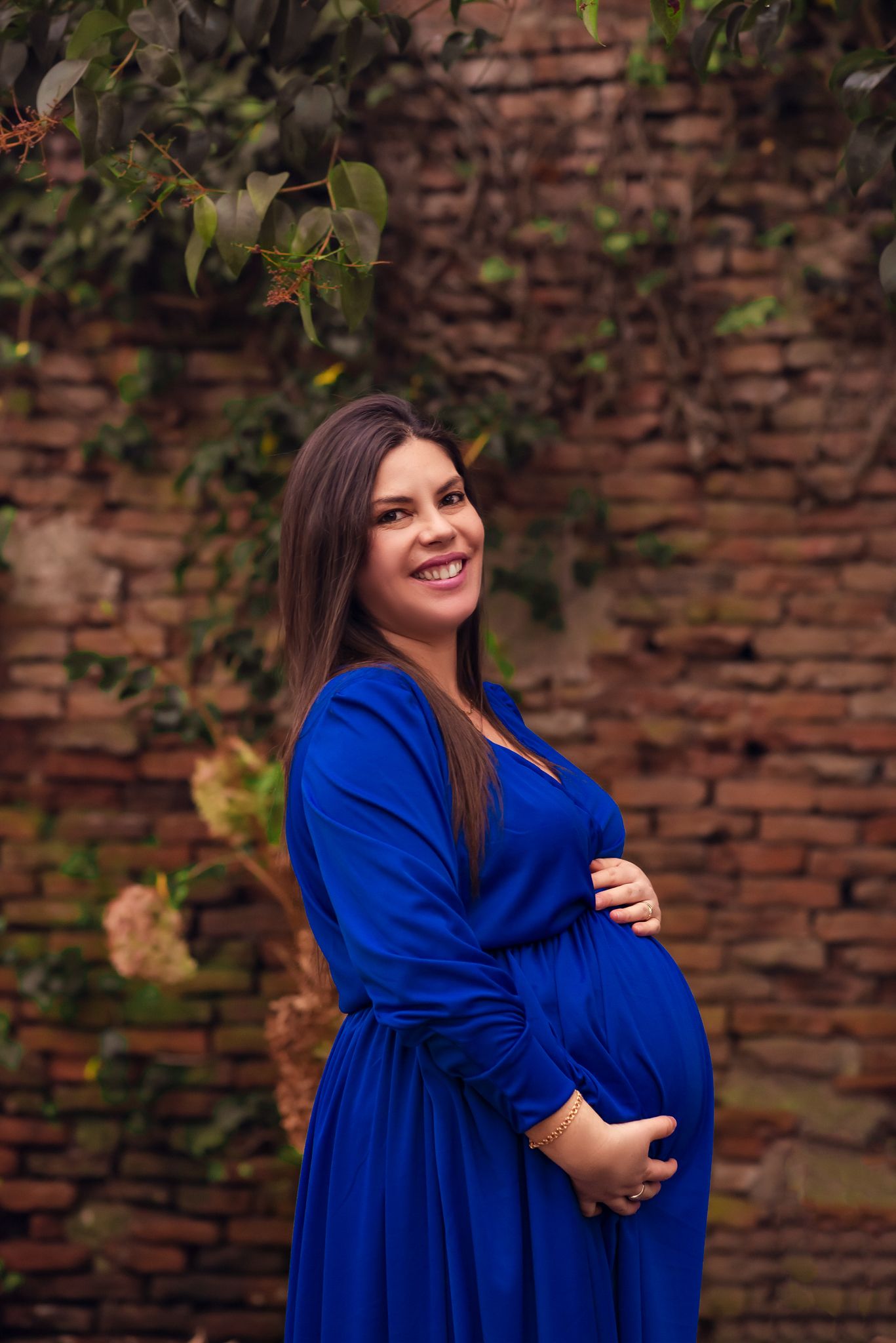 A pregnant woman in a vibrant royal blue dress poses outdoors against a rustic brick wall with greenery.