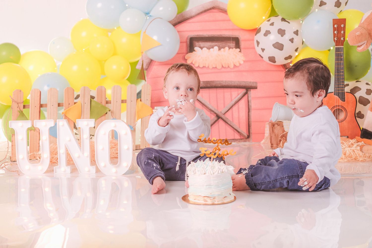 Two small children sit on the floor at a birthday party with a cake and yellow and pink decorations.