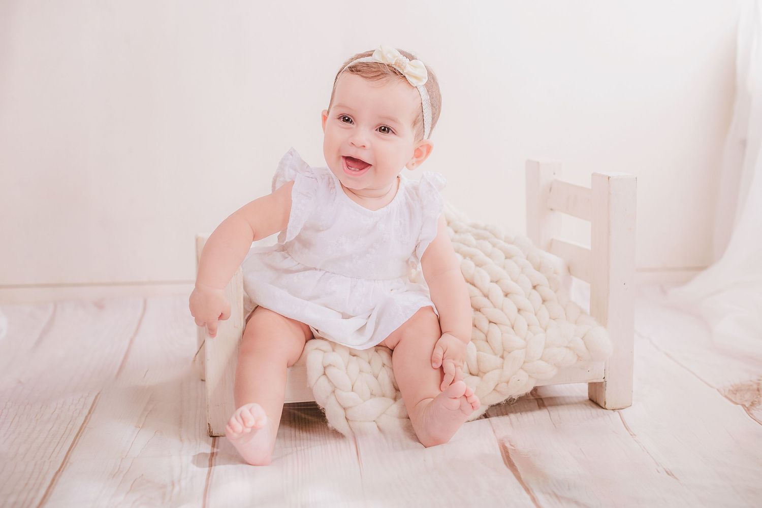 A small child in a white dress sits on a wooden floor with a cream-colored knit blanket.