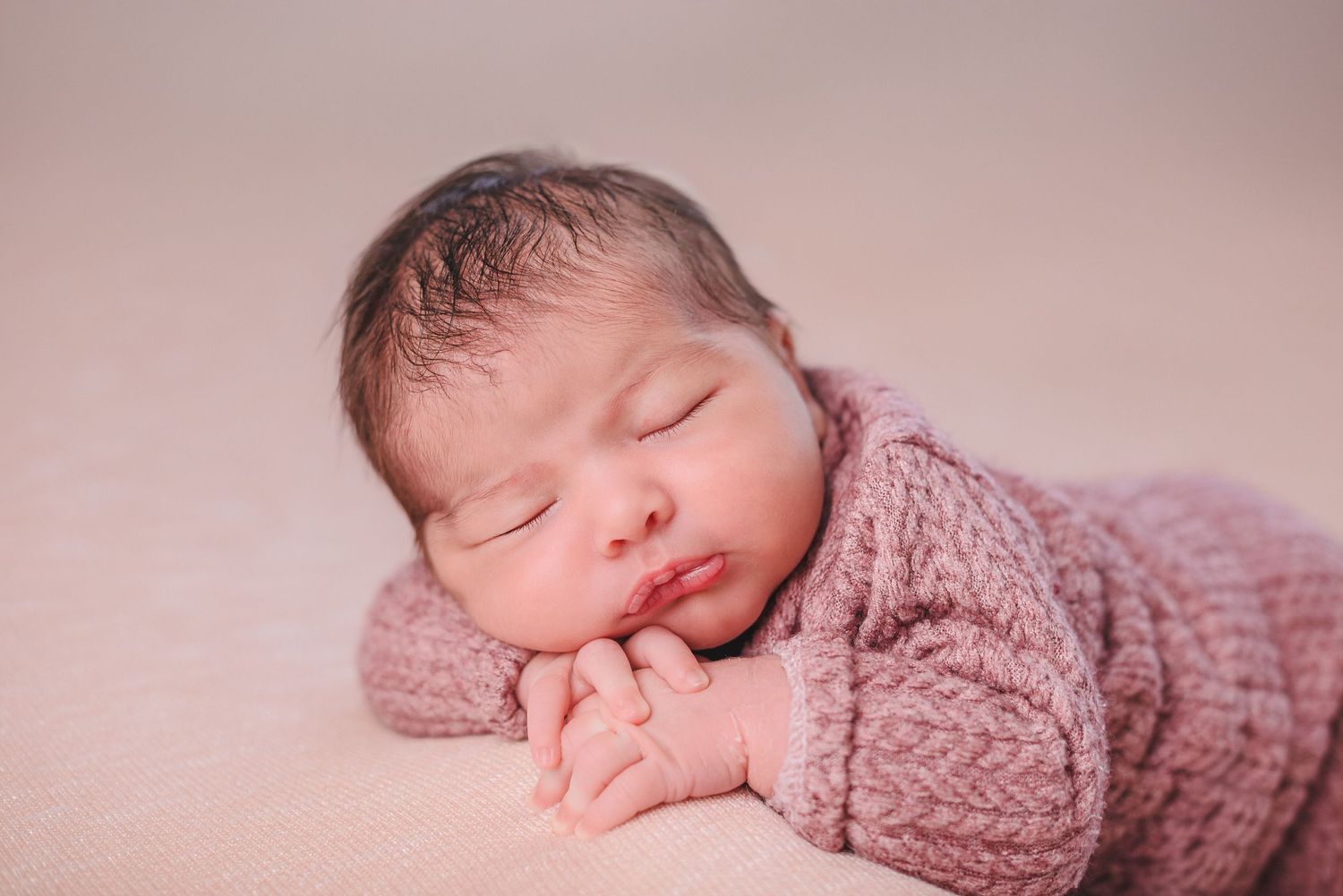 Newborn baby sleeping peacefully in soft pink knitted wrap during professional photo session.
