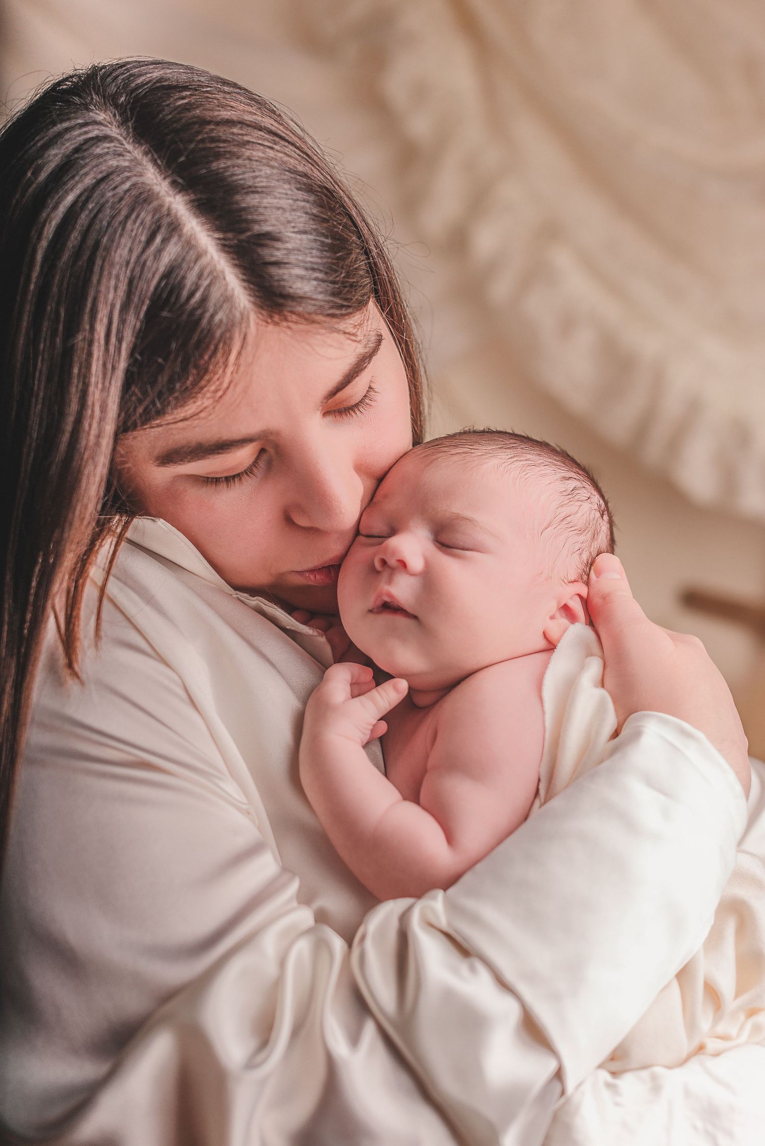 A tender moment captured as a mother in a white sweater gently kisses her sleeping newborn baby.