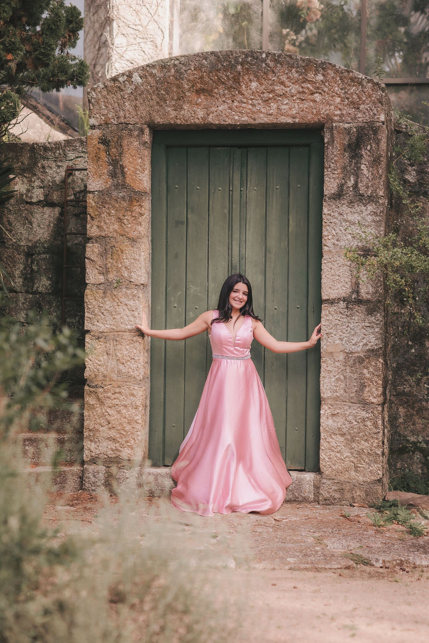 A pink formal dress is showcased against rustic stone walls and green wooden doors in a photo series.