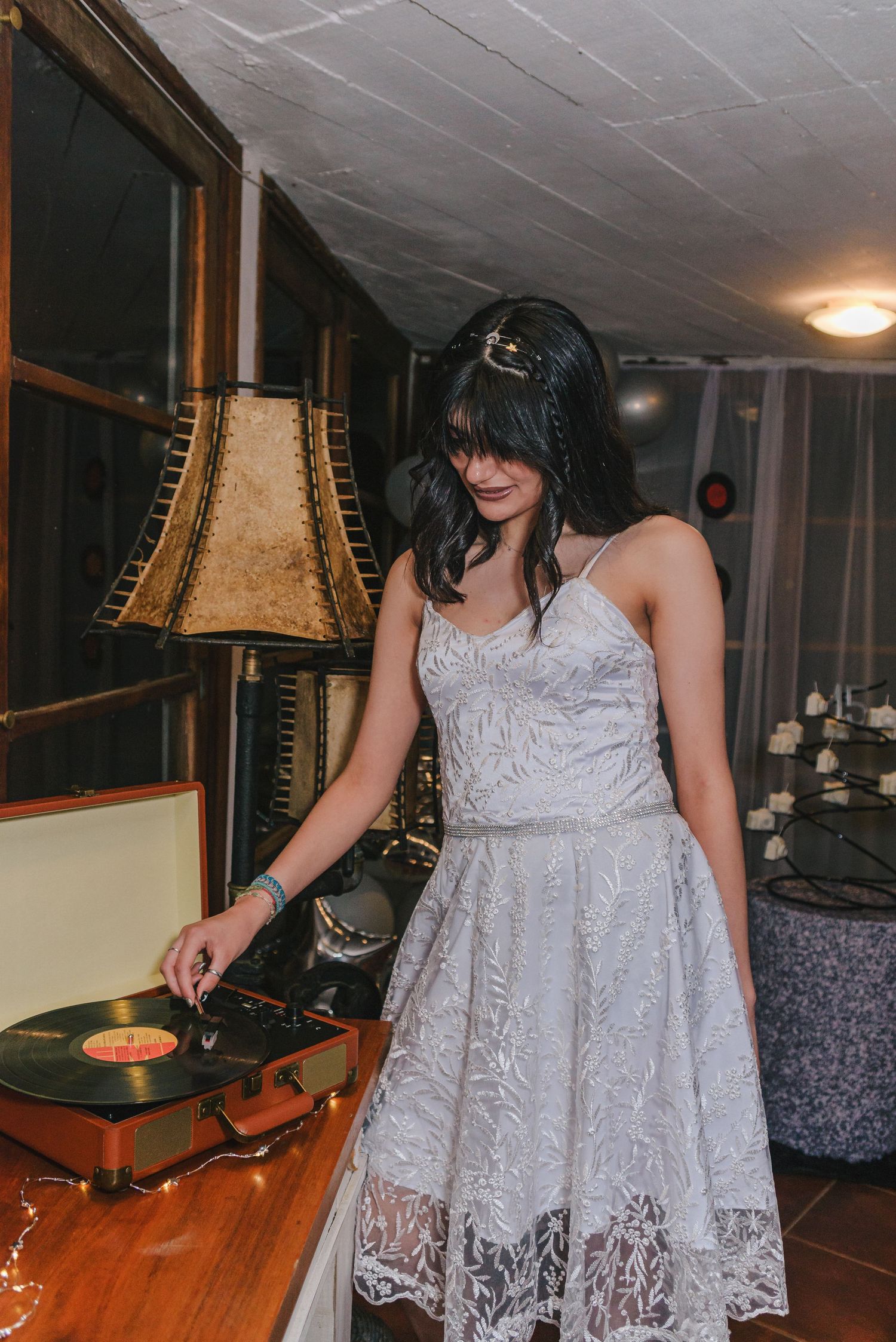 A person in a white lace dress stands in a dimly lit room near vintage-style table lamps.