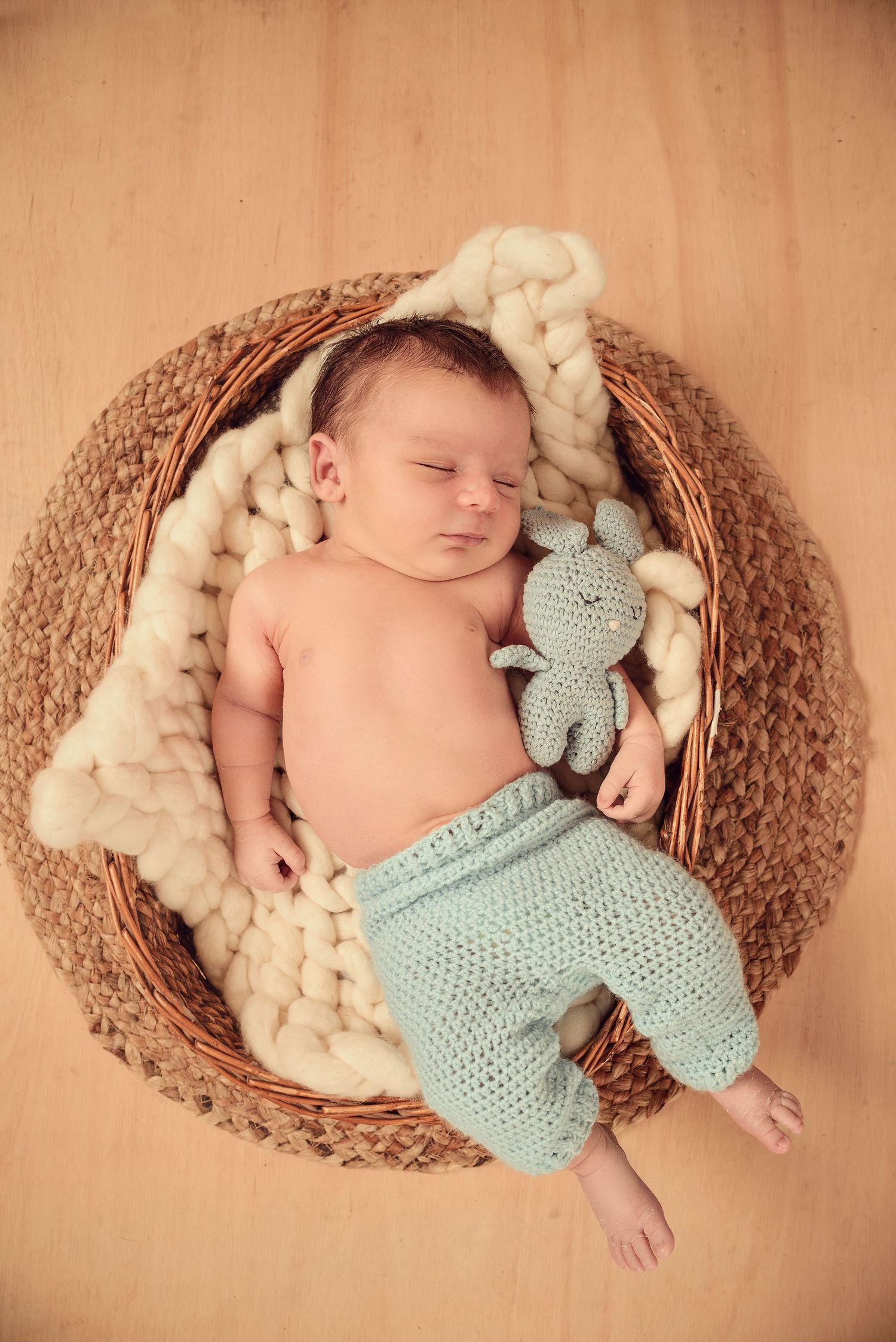 Newborn baby sleeping peacefully in brown basket with cream wool blanket and blue knitted teddy bear companion.
