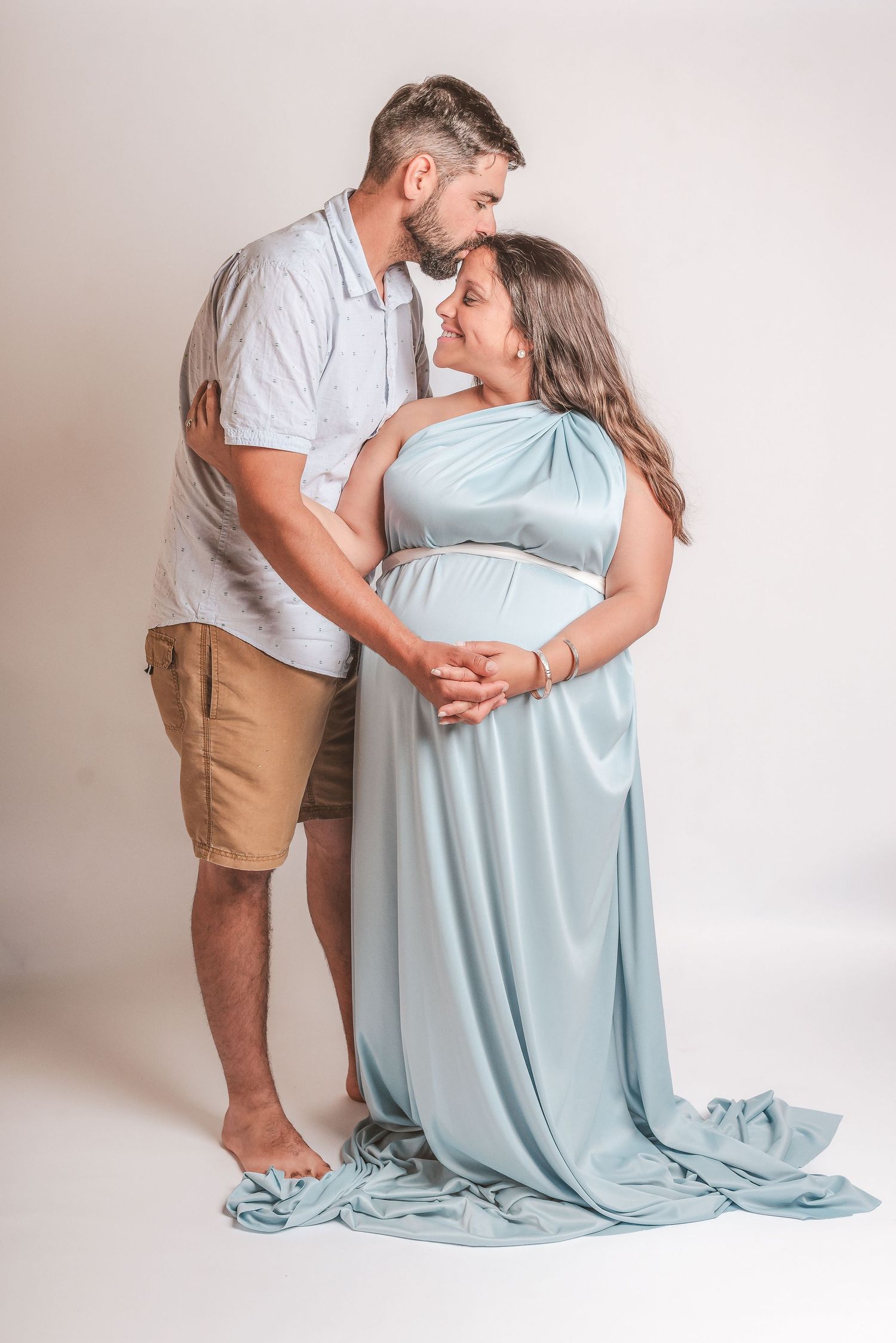 A couple in soft lighting poses for a romantic maternity photo series wearing light blue and beige against a white backdrop.