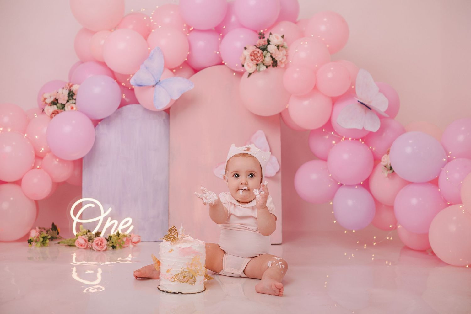 Pink and lavender balloon arch decorates a first birthday cake smash photo setup with floral accents against pink backdrop.