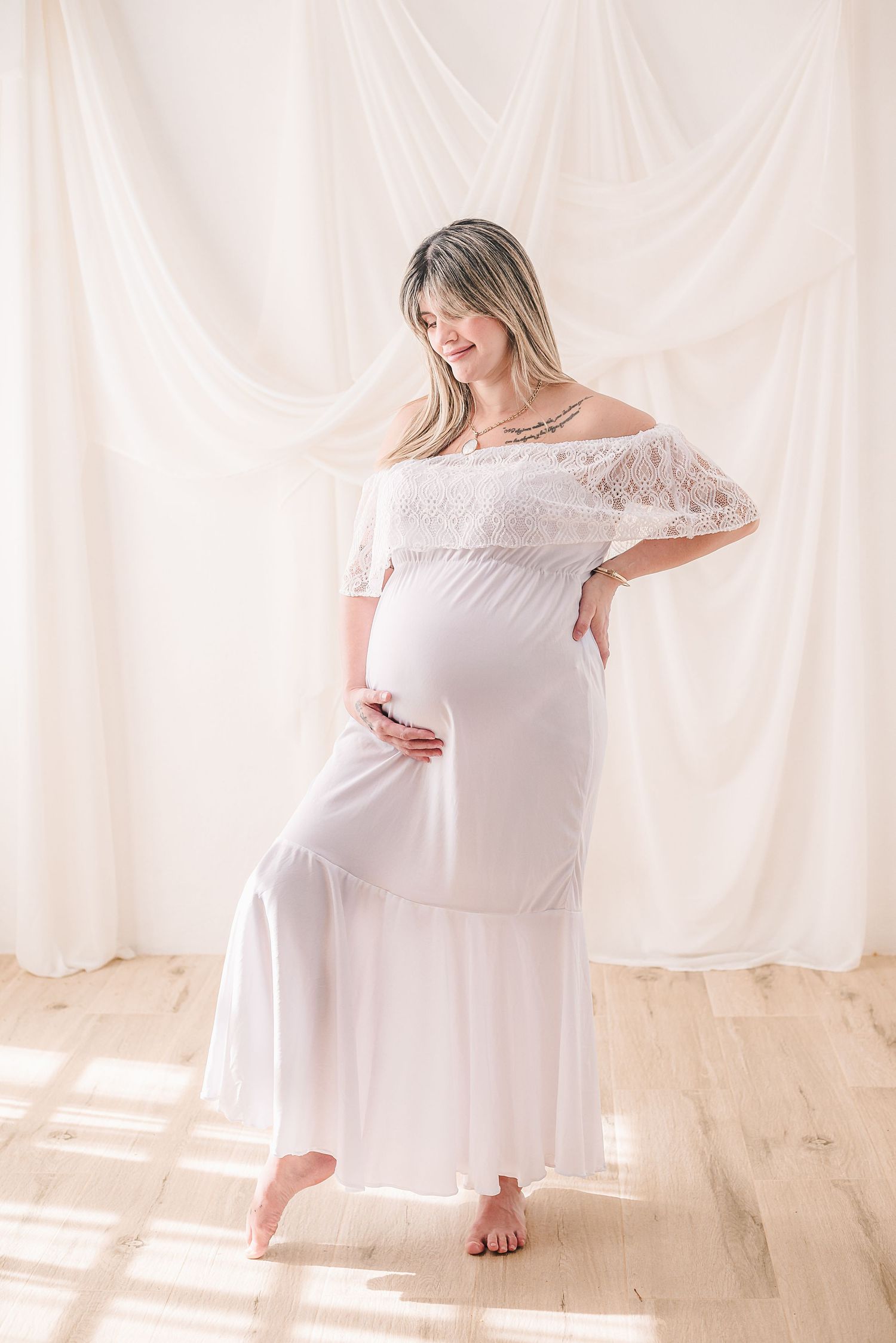 A maternity photo series showing an expectant mother in a white off-shoulder dress posing against flowing white curtains.