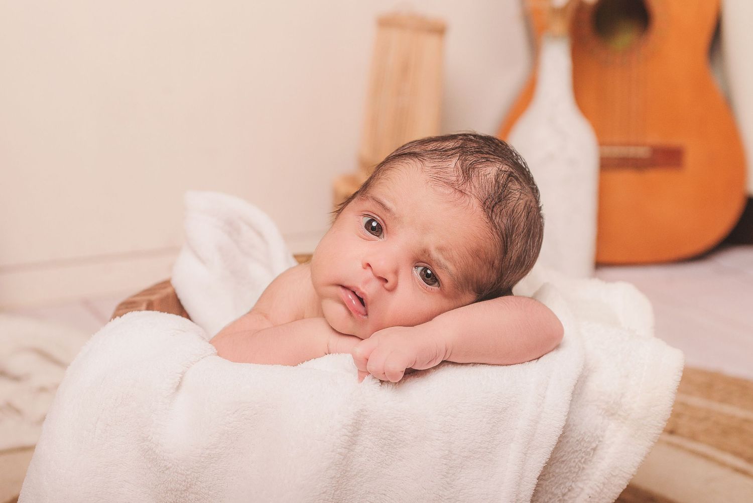 A small infant rests on a soft white blanket with a guitar visible in the background.