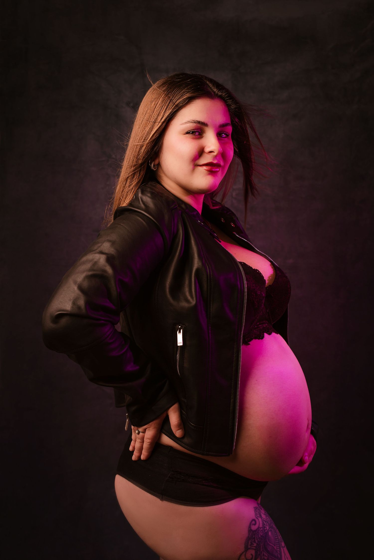 A maternity photoshoot featuring dramatic lighting with pink and purple hues against a dark background.