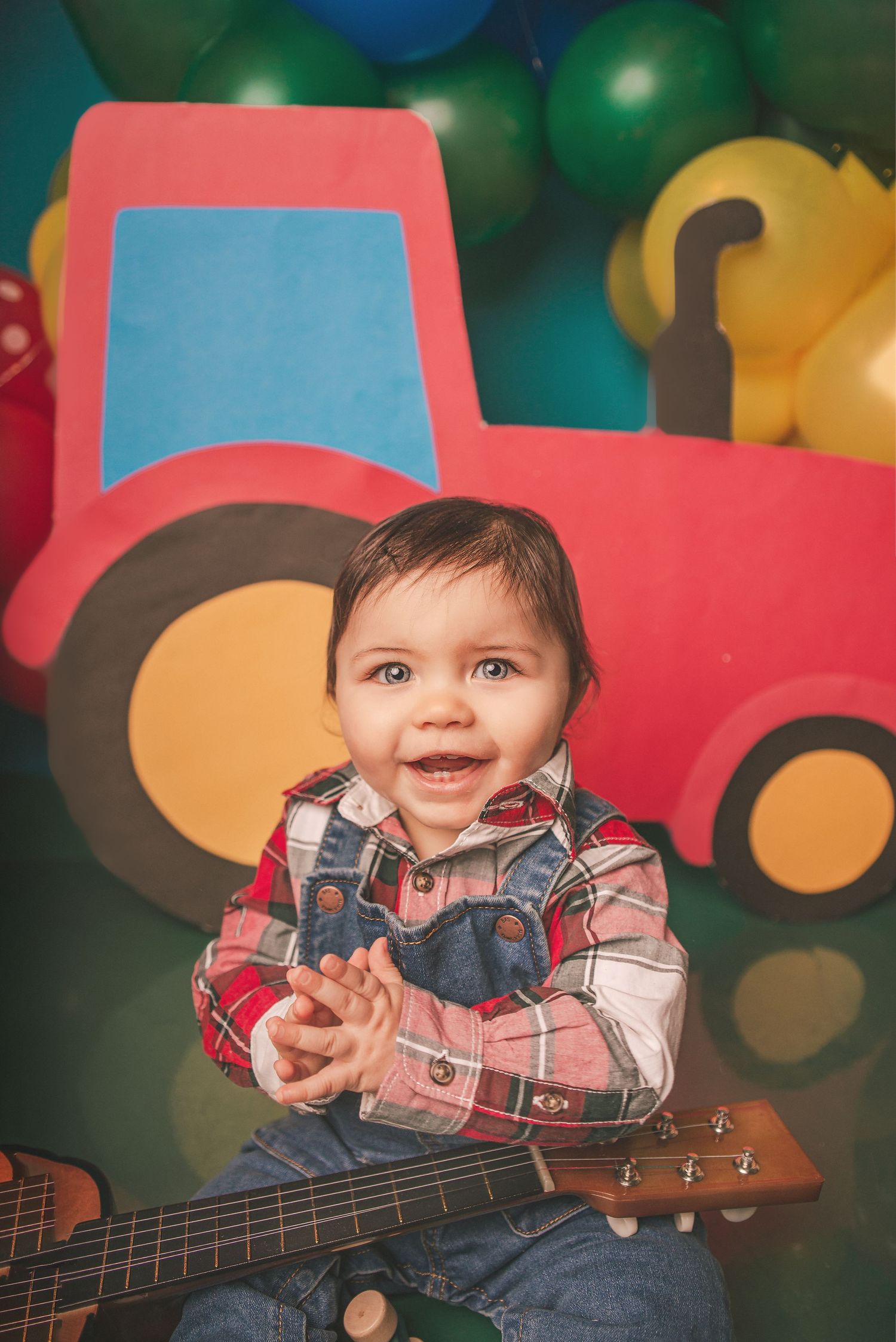 A child in overalls and plaid shirt poses with a ukulele against a colorful bokeh background with balloons.